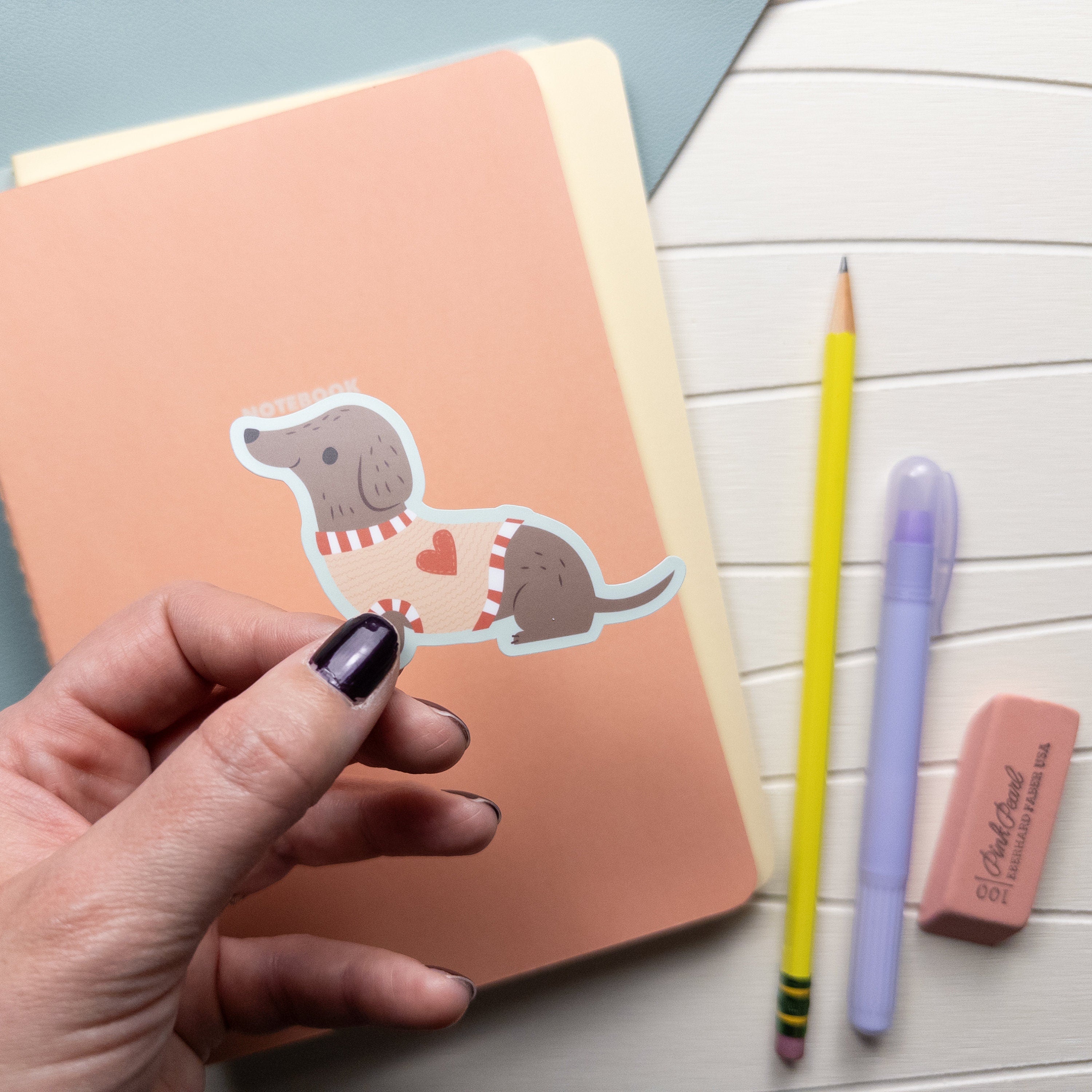 Hand holding an illustrated waterproof vinyl sticker of a dachshund wearing a sweater with a heart on it.