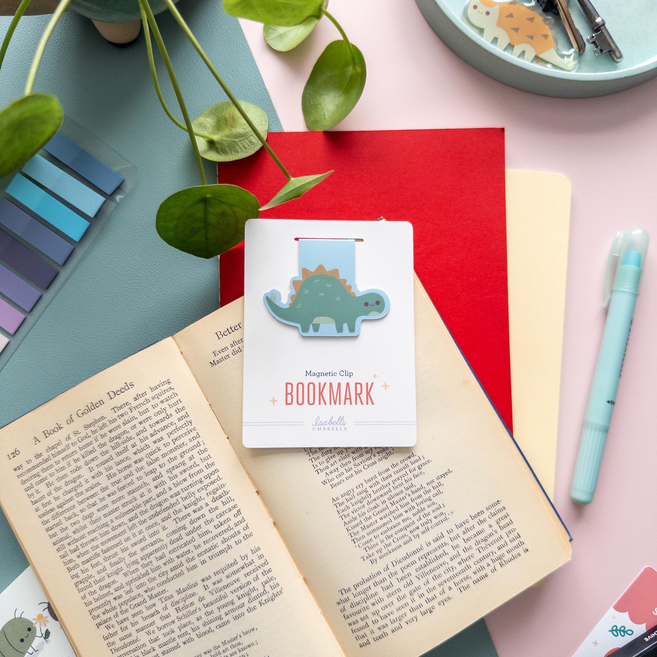 Cute Stegosaurus magnetic bookmark with matte lamination, kawaii dinosaur clip for books, journals, and planners. Pictured laying on top of an open book.