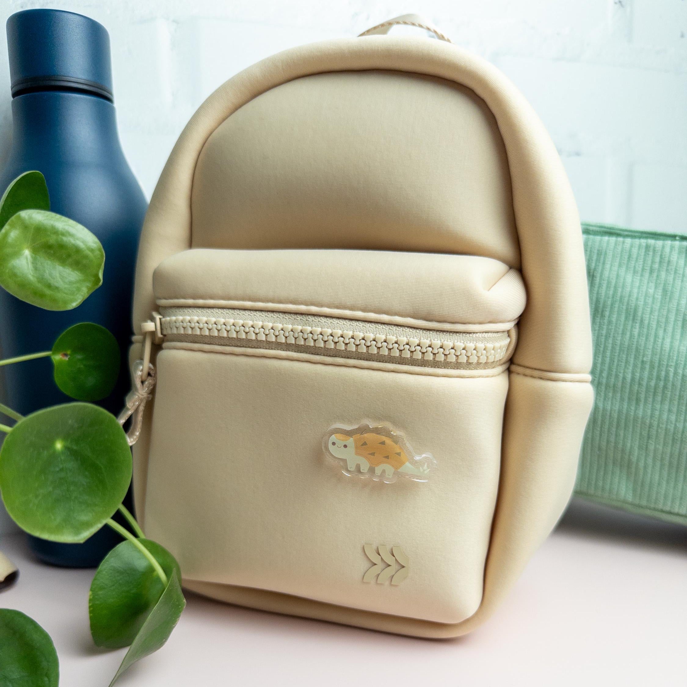 Adorable acrylic pin featuring a chubby ankylosaurus made from durable acrylic and finished with a smooth epoxy finish. The pin is attached to a beige backpack and is pictured with a blue water bottle, plant, and sage green pouch.
