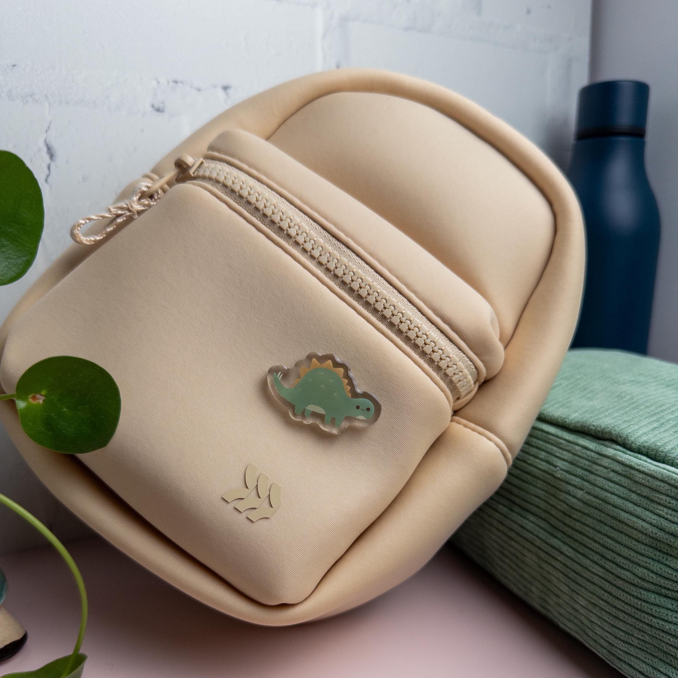 A cute green Stegosaurus acrylic pin with glossy epoxy finish attached to a beige backpack. It is photographed with a blue water bottle in the background 