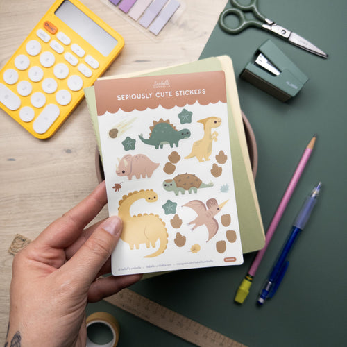 Hand holding a sheet of stickers featuring six different dinosaurs, foot prints, and leaves.