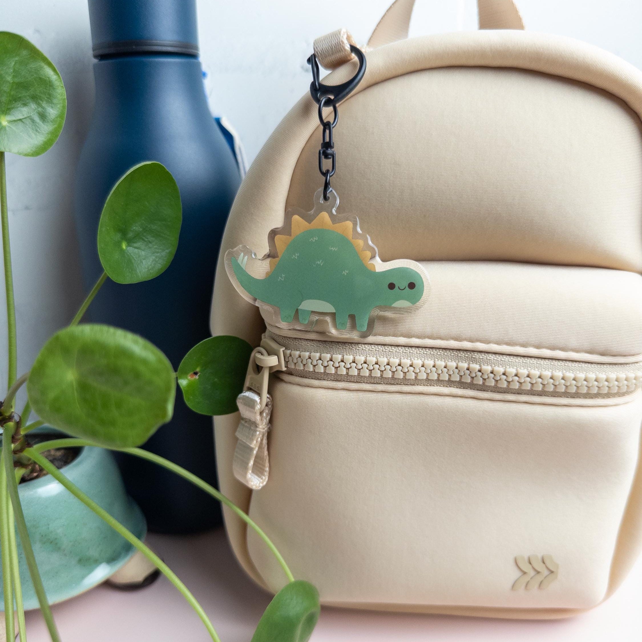 Cute Stegosaurus acrylic keychain with glossy epoxy finish hanging from a zipper on a cream colored backpack.