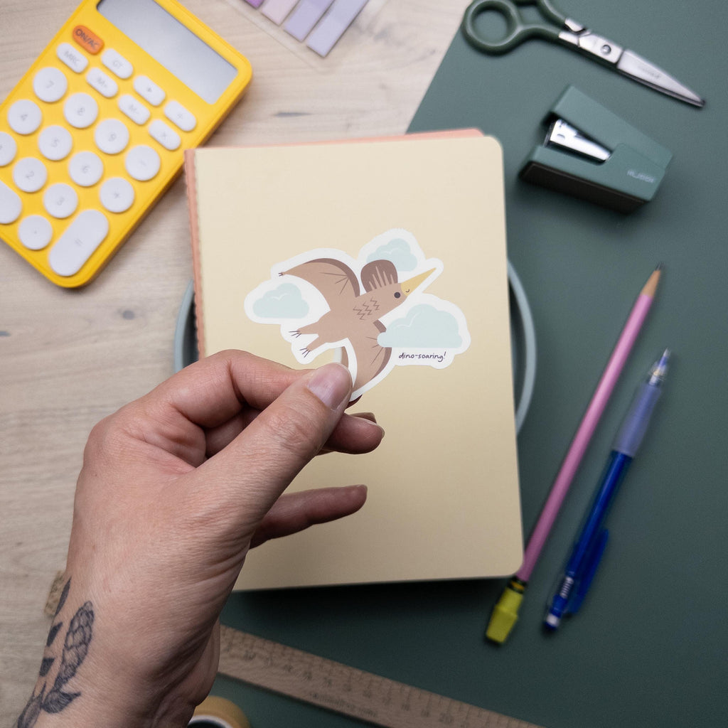 Hand holding a cute illustrated pterodactyl vinyl sticker, waterproof flying dinosaur decal for laptops, water bottles, and journals, kawaii prehistoric design.