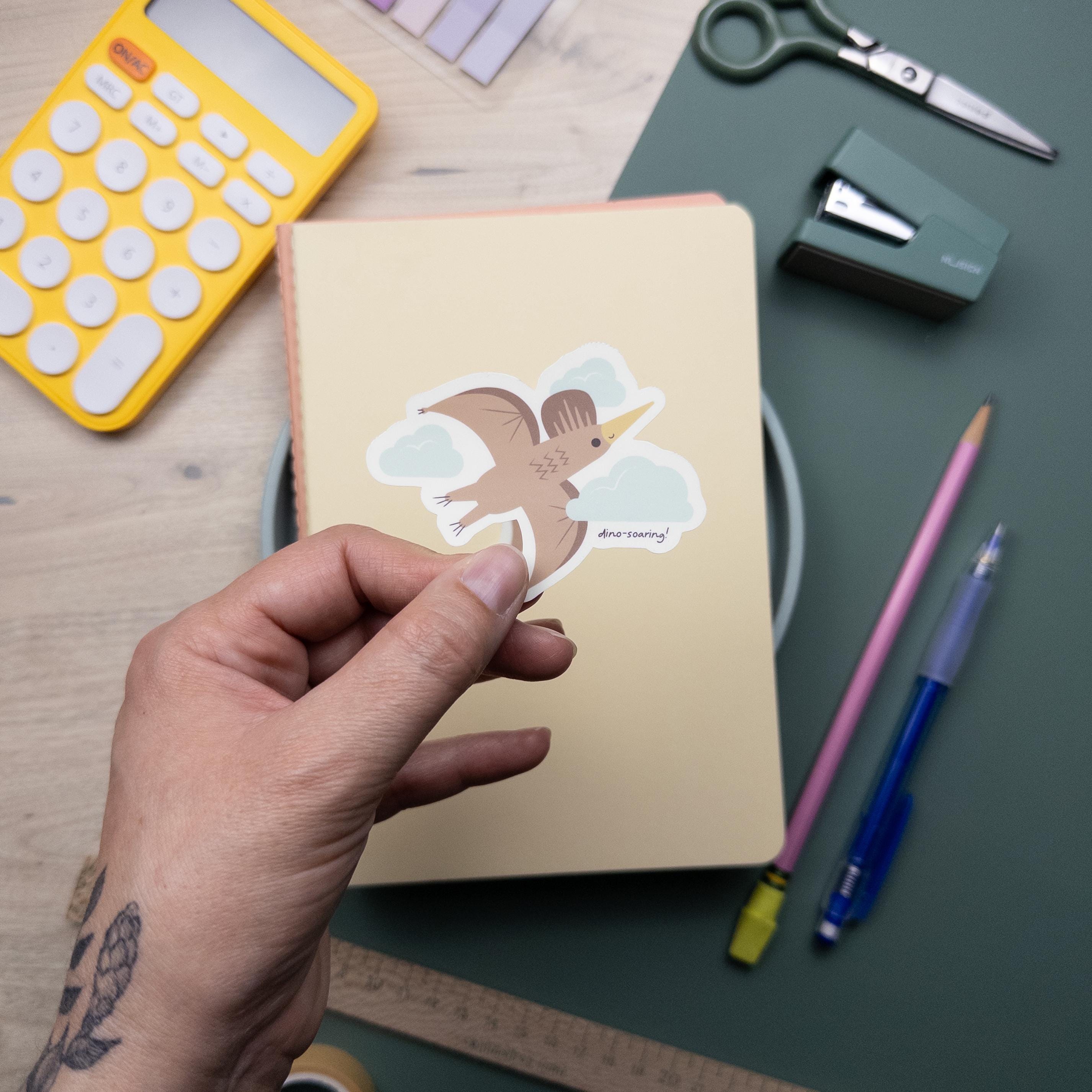 Hand holding a cute illustrated pterodactyl vinyl sticker, waterproof flying dinosaur decal for laptops, water bottles, and journals, kawaii prehistoric design.