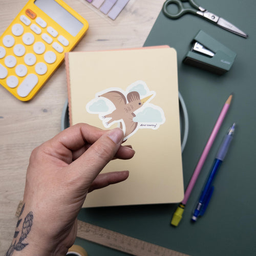 Hand holding a cute illustrated pterodactyl vinyl sticker, waterproof flying dinosaur decal for laptops, water bottles, and journals, kawaii prehistoric design.