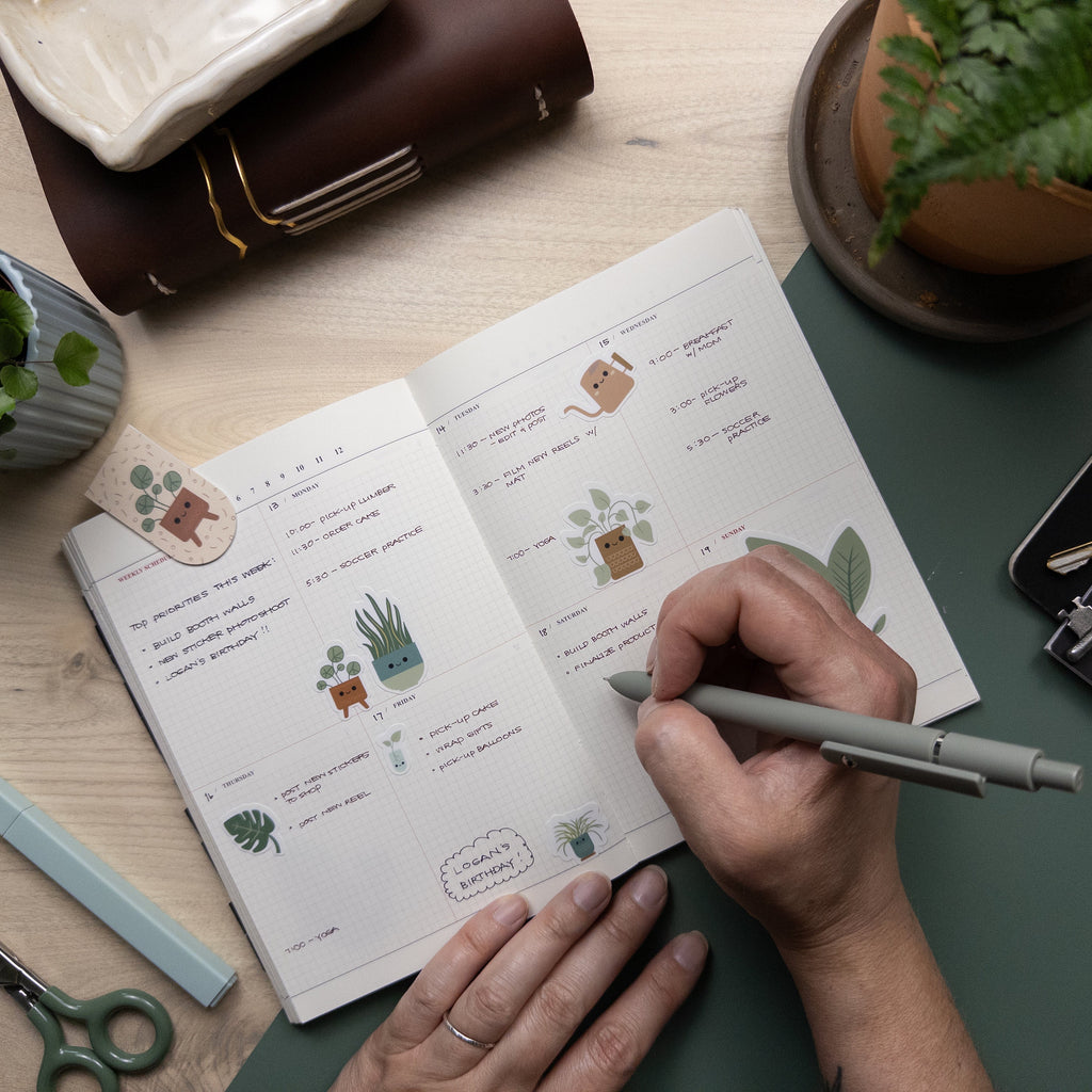 Hand writing in weekly planner that has been decorated with stickers of potted house plants.