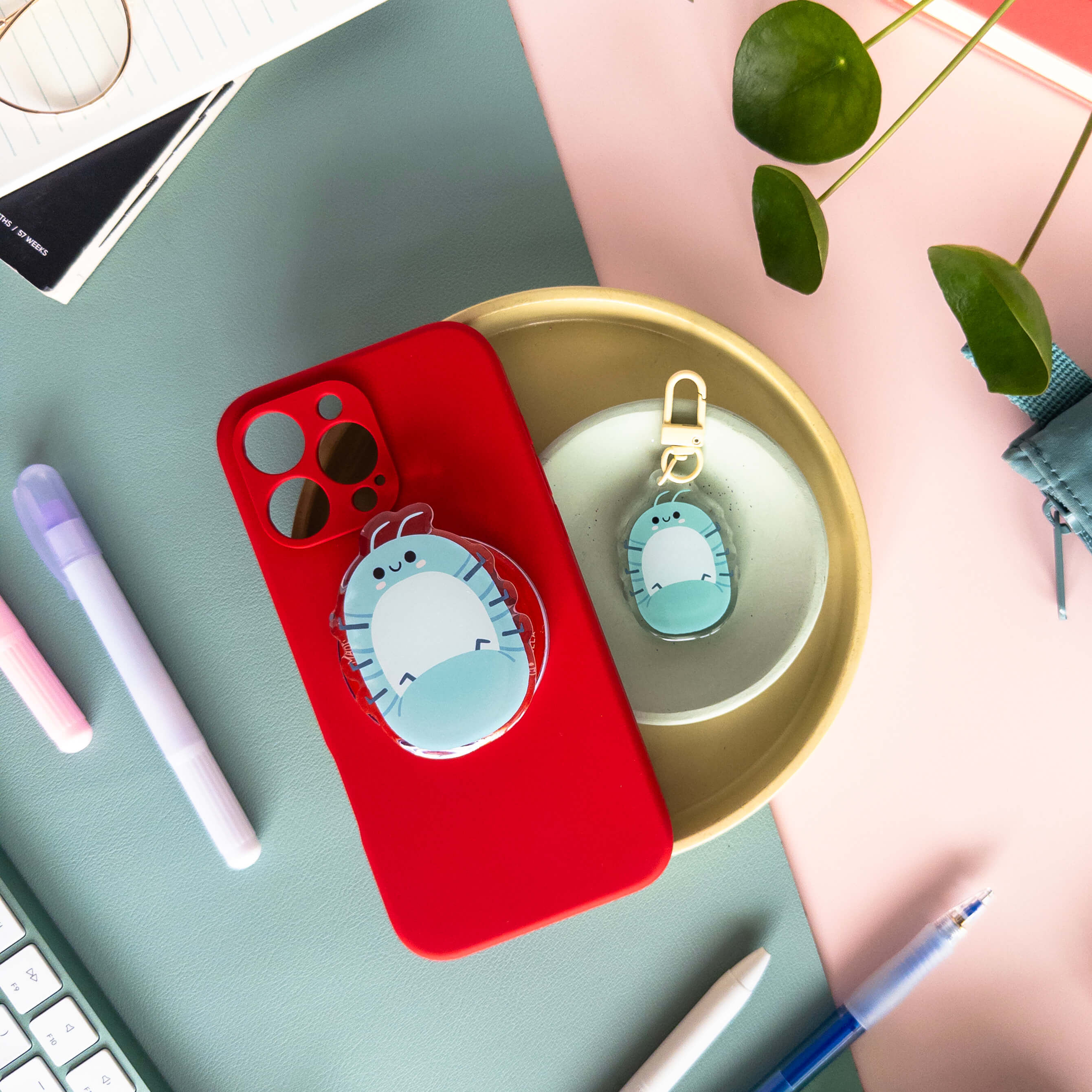 Red phone case with a blue roly poly magnetic phone grip with roly poly keychain charm on a desk with pens and a plant.