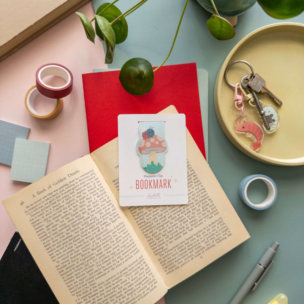 Open book with a magnetic bookmark of a mushroom with a ladybug on top, tape and keychain on a table