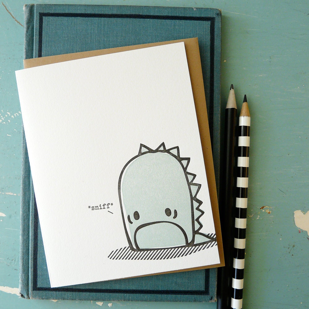 A cute sad monster illustrated letterpress card set on top of a book with two pencils beside it.