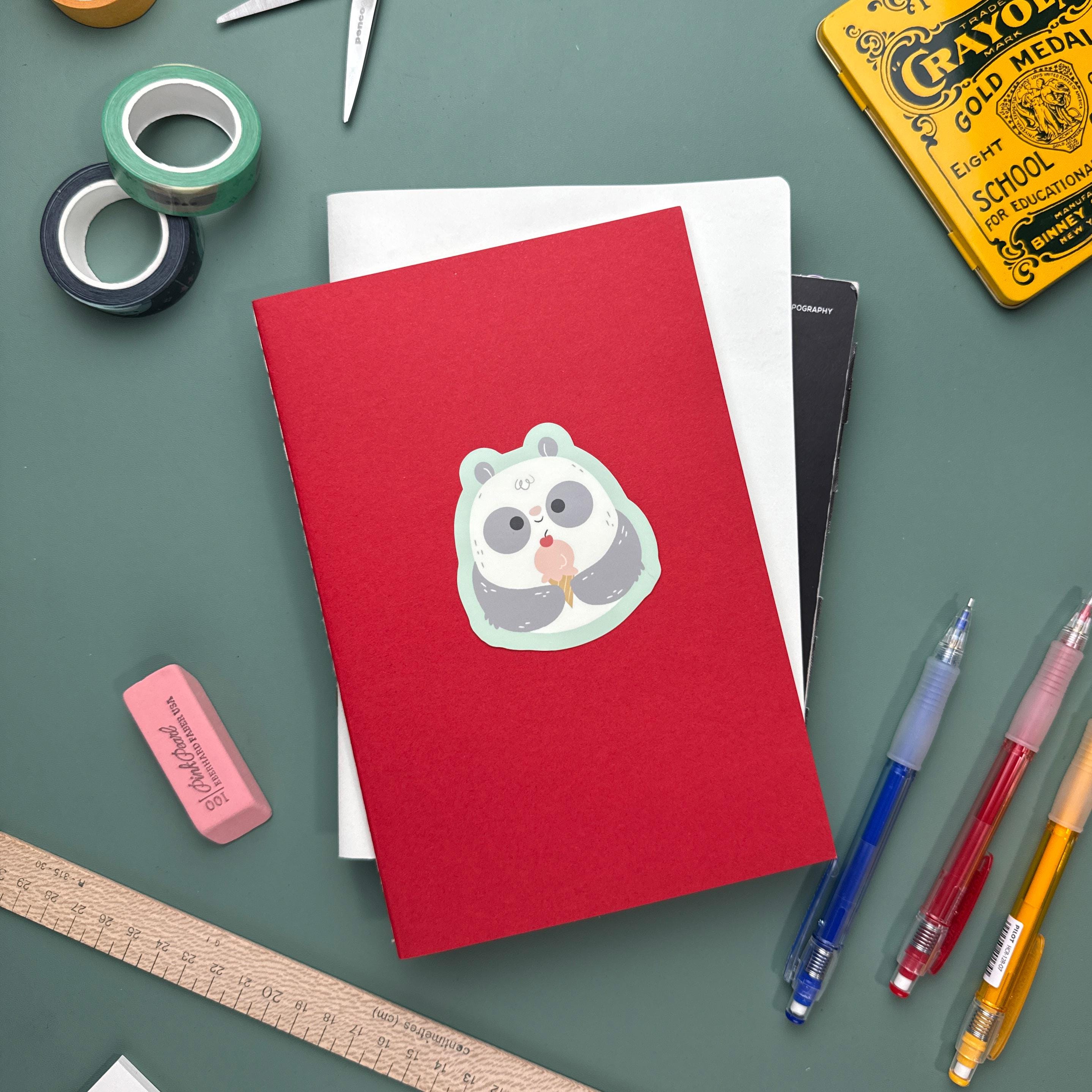 A red notebook with a panda ice cream sticker placed in the center lies on top of a white notebook. Surrounding items include pens, a ruler, an eraser, scissors, washi tape, and a vintage yellow Crayola tin.