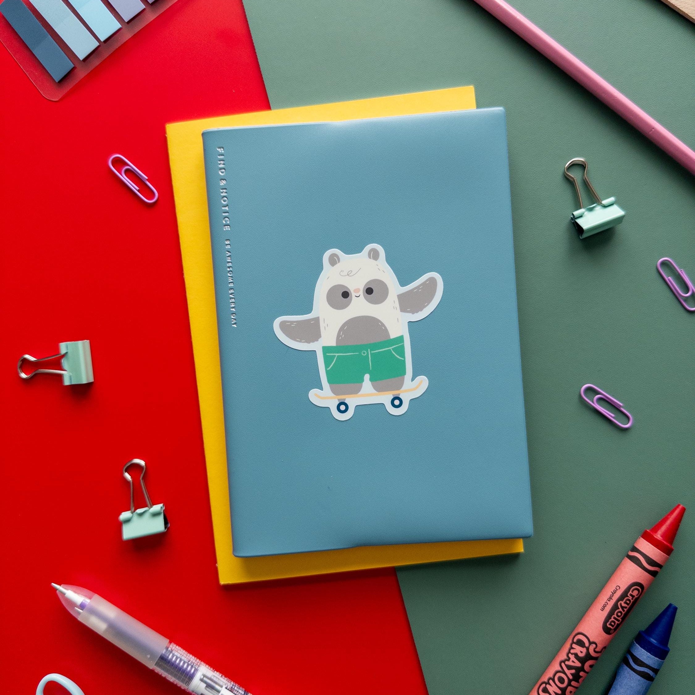 Cute skateboarding panda vinyl sticker sitting on top of a blue and yellow notebooks with a variety of art supplies surrounding it.