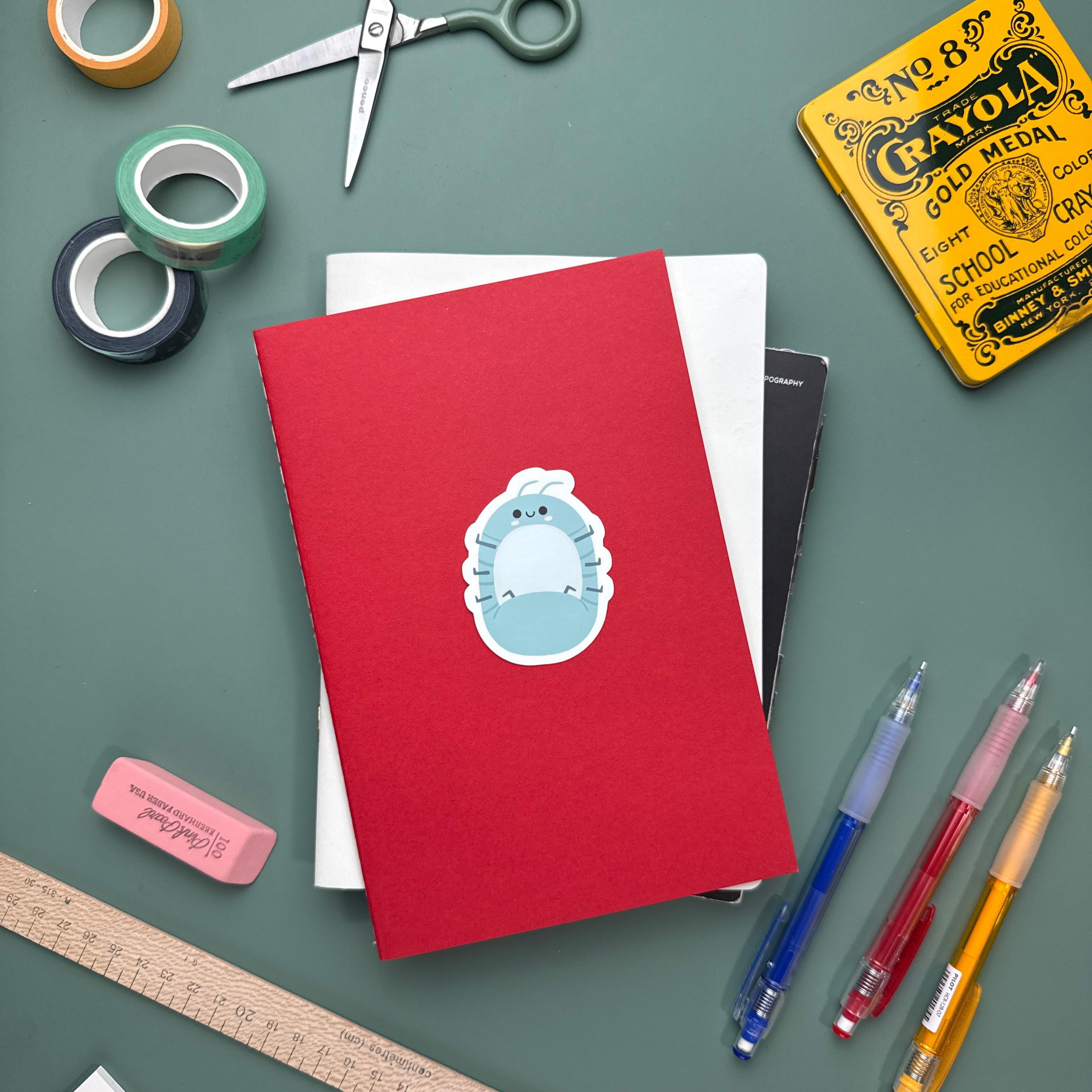 Kawaii roly poly sticker applied to the cover of a red notebook. The scene includes colorful pens, a ruler, and other stationery, highlight this cute bug sticker as a fun addition to planners or journals.