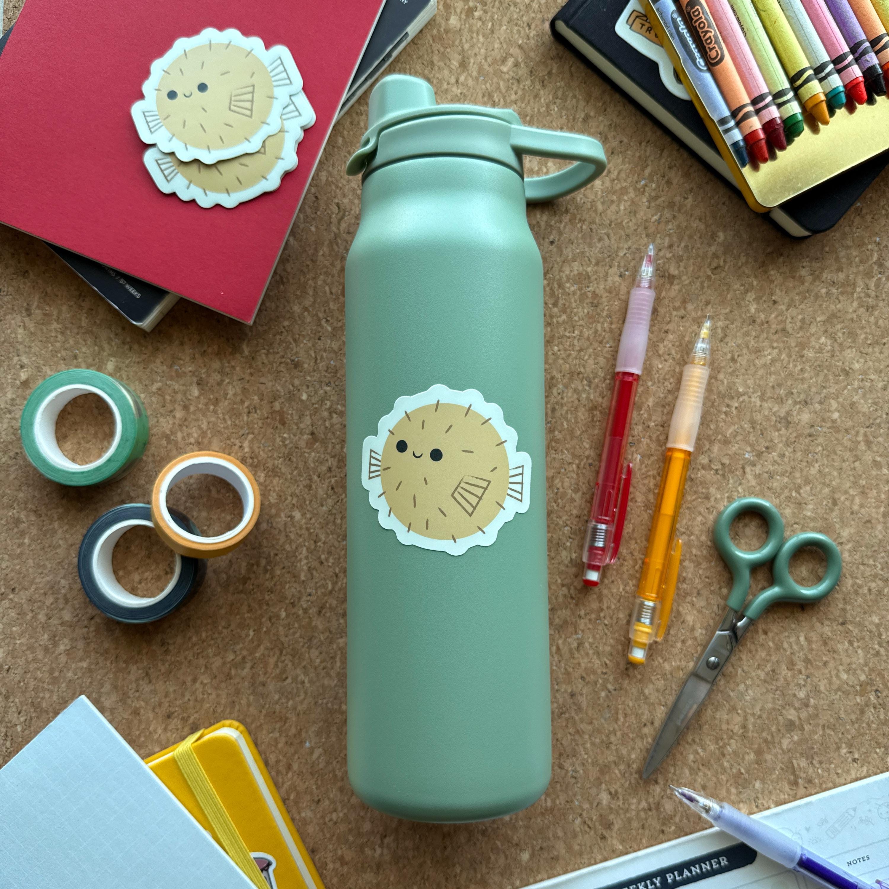 Cute puffer fish sticker on a sage green water bottle, surrounded by colorful pens, washi tape, and notebooks. This waterproof vinyl sticker is perfect for decorating bottles, laptops, and school supplies.