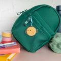 Small green backpack with a yellow  decorative pufferfish charm on a surface with stationery items