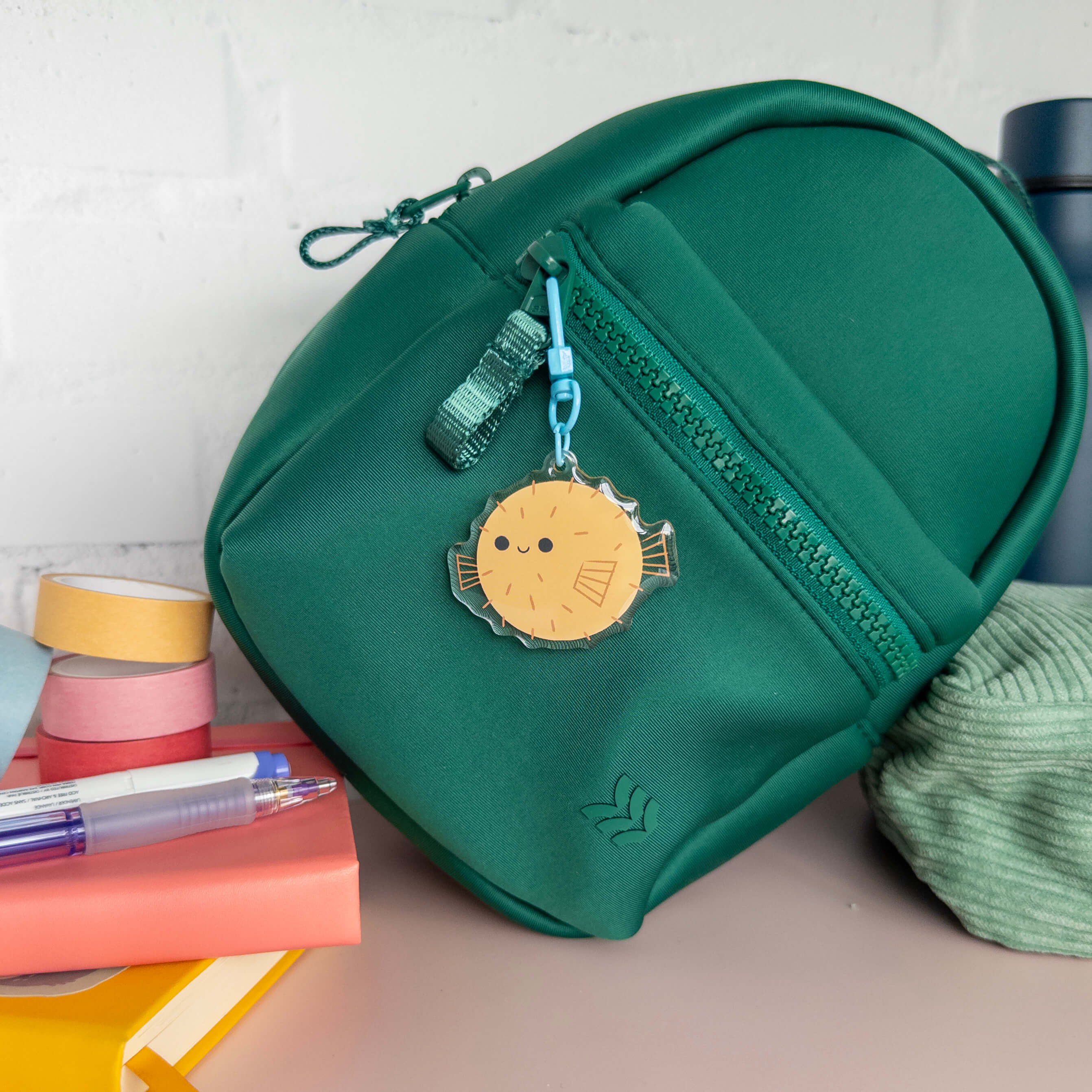Small green backpack with a yellow  decorative pufferfish charm on a surface with stationery items