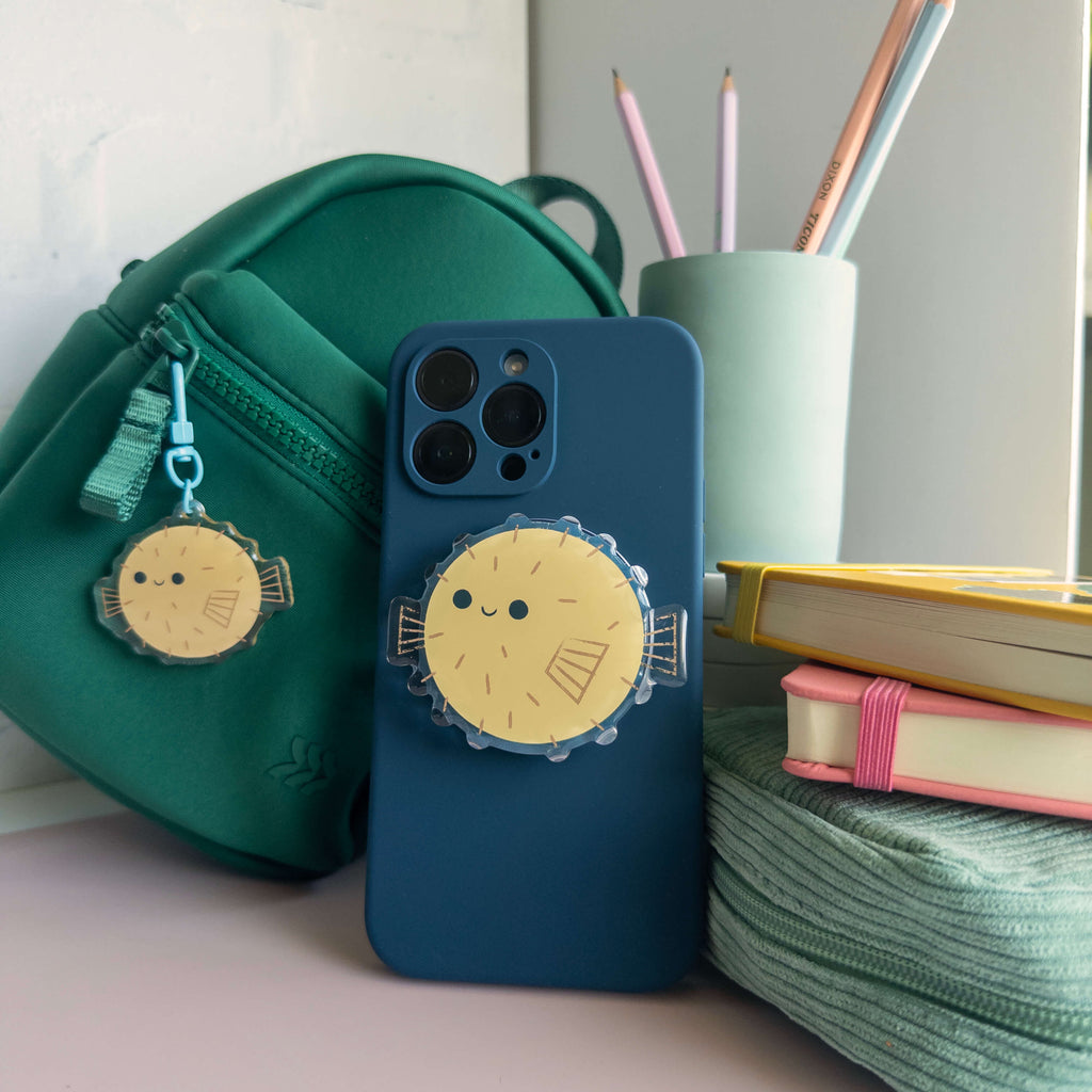 Blue phone case with pufferfish designed magnetic phone grip next to a green bag and stationery items on a desk.