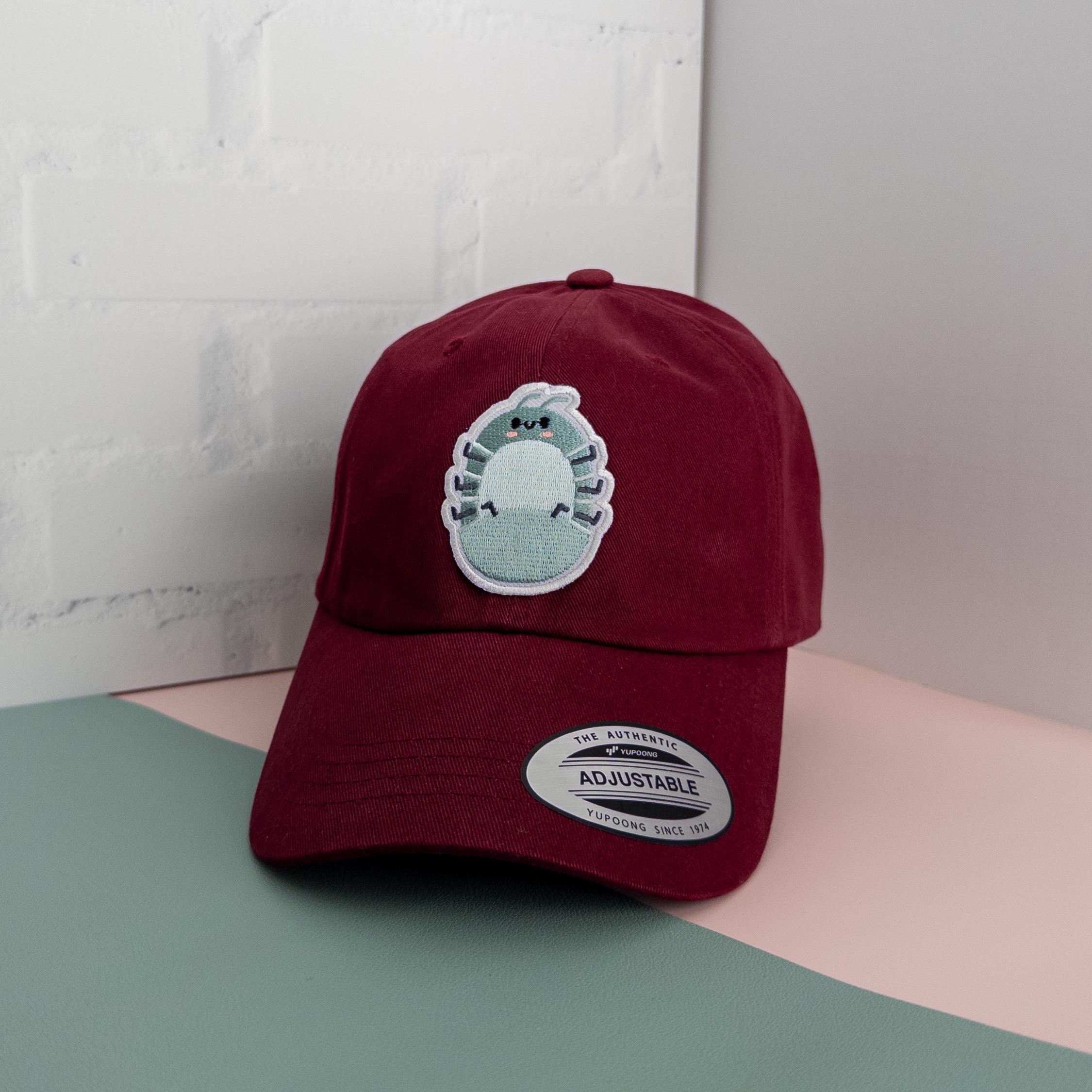 A maroon cotton twill dad hat with embroidered roly poly patch sitting on top of a pink and sage green table