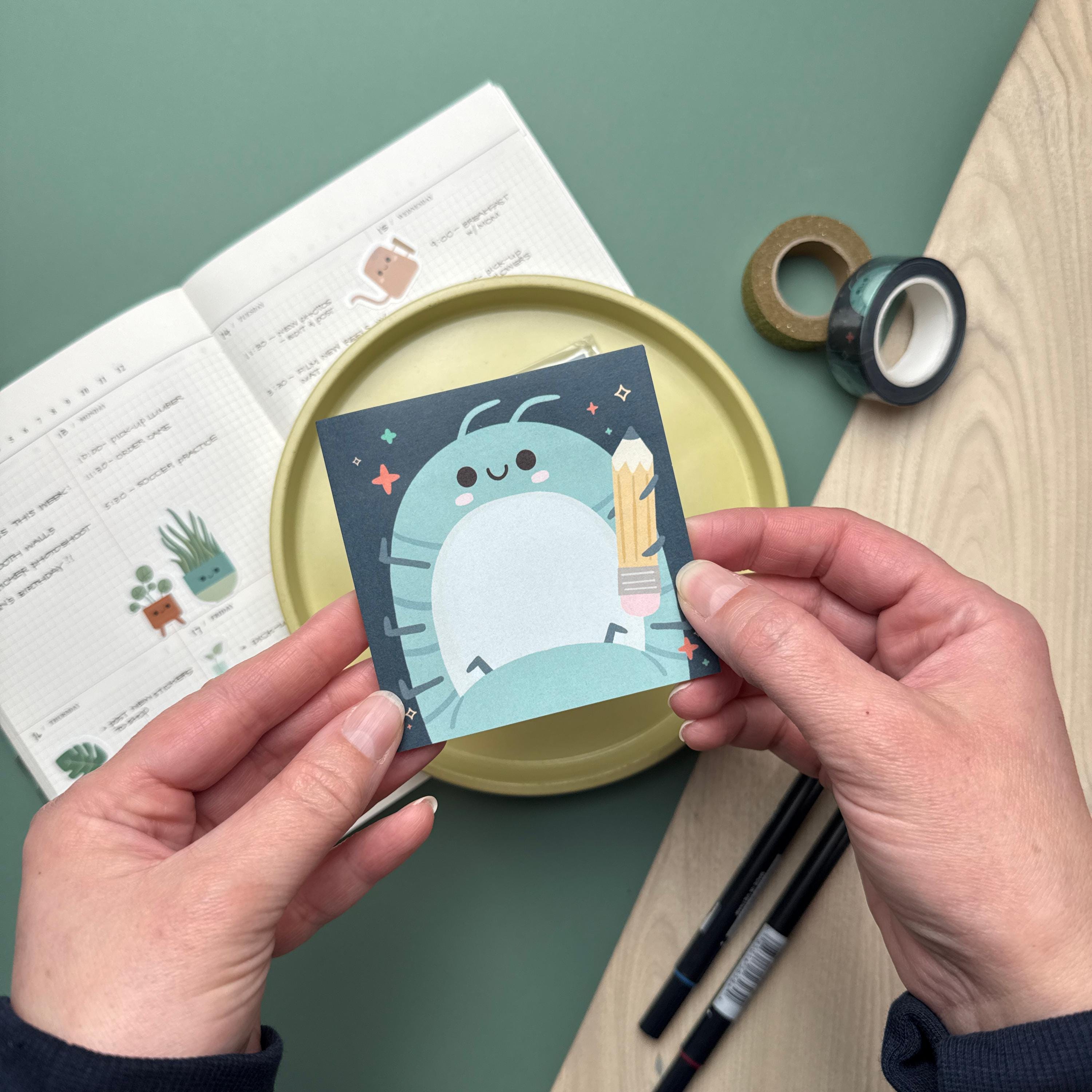 Hands holding a sticky notepad featuring a roly poly holding a pencil. The notepad is being held above a yellow plate and is surrounded by a calendar, markers, ruler, and another notepad.