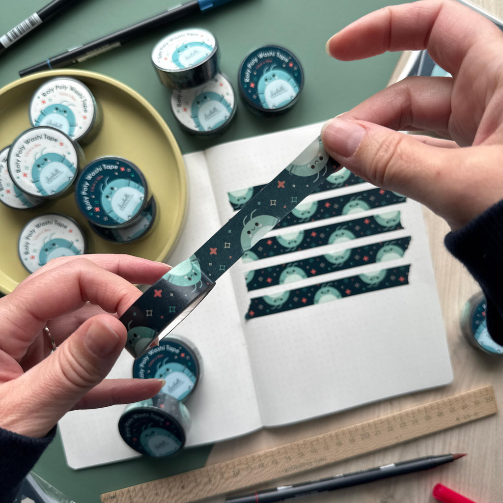 A hand holding a roll of Roly Poly-themed Washi tape with a dark background, featuring a smiling isopod illustration. More rolls of the same Washi tape and an opened journal decorated with strips of the tape are visible in the background.