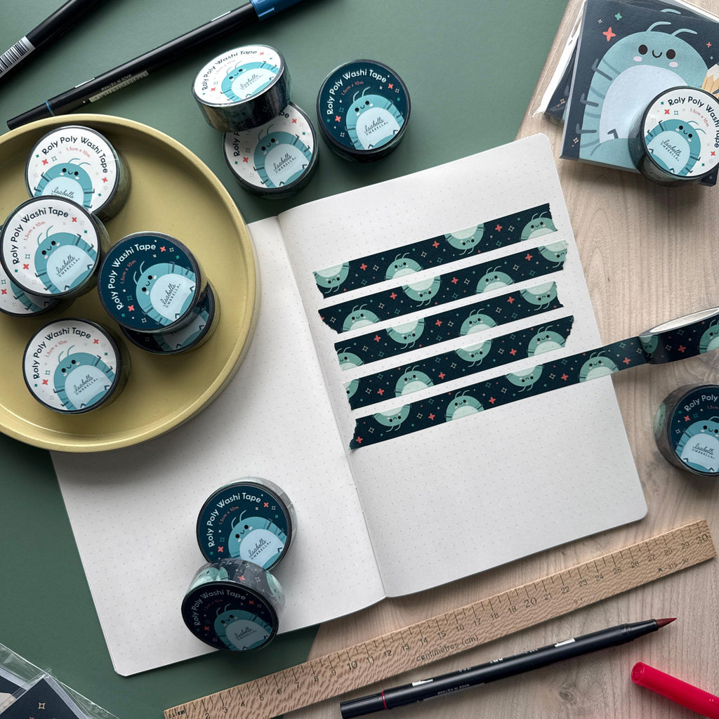 Multiple rolls of Roly Poly-themed Washi tape are arranged on a yellow tray and around an opened journal. The journal showcases evenly spaced strips of the dark Washi tape, adorned with a smiling isopod pattern.