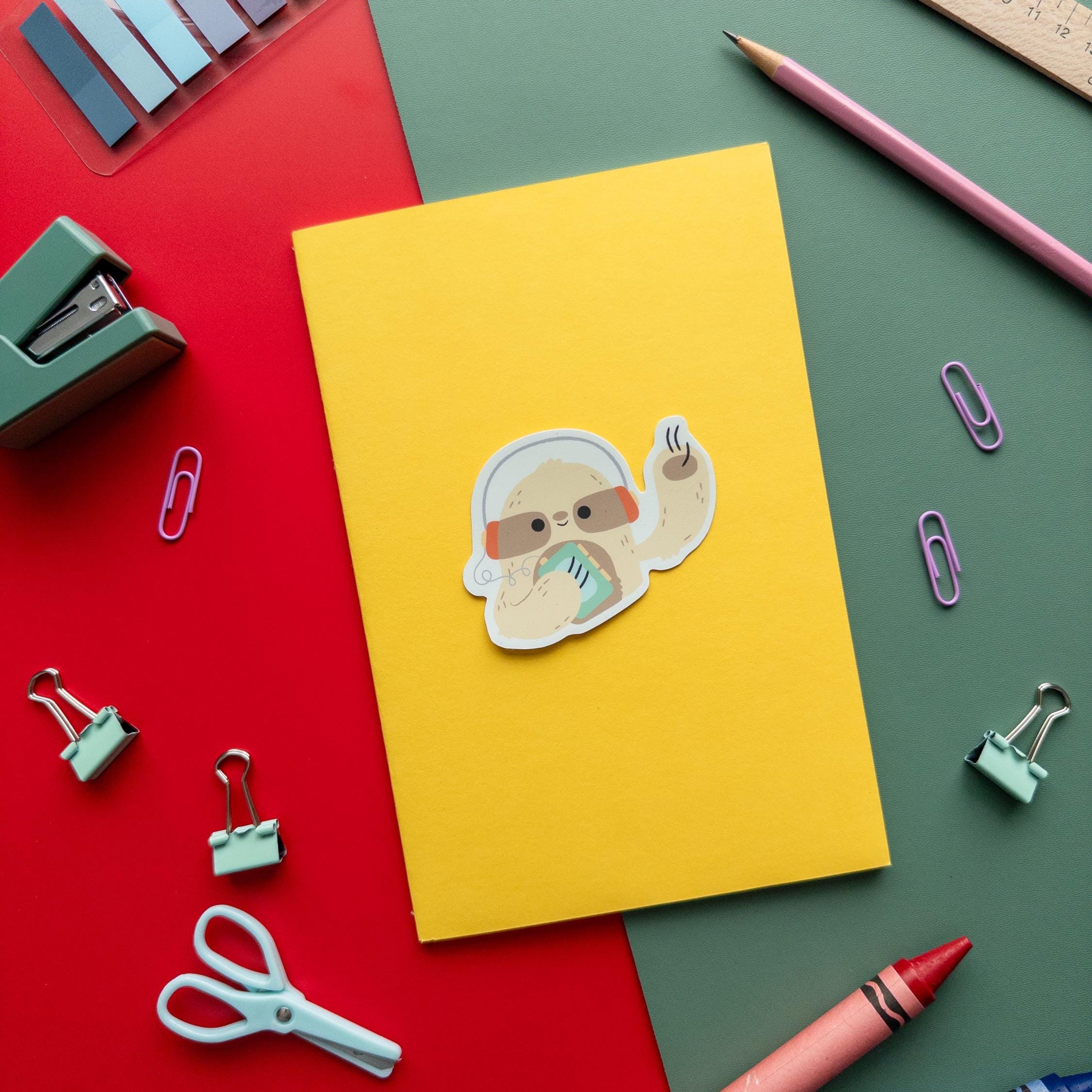 Adorable sloth wearing a walkman sticker photographed on top of a yellow notebook with a sticker, scissors, crayons, and a ruler on a red and green background.