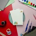 Flat lay of the snail and bee-themed notepad from Isabell’s Umbrella on a colorful desk setup. Surrounding items include watercolor paints, paintbrush, pens, washi tape, a ruler, and a pencil case, creating a cozy stationery scene.