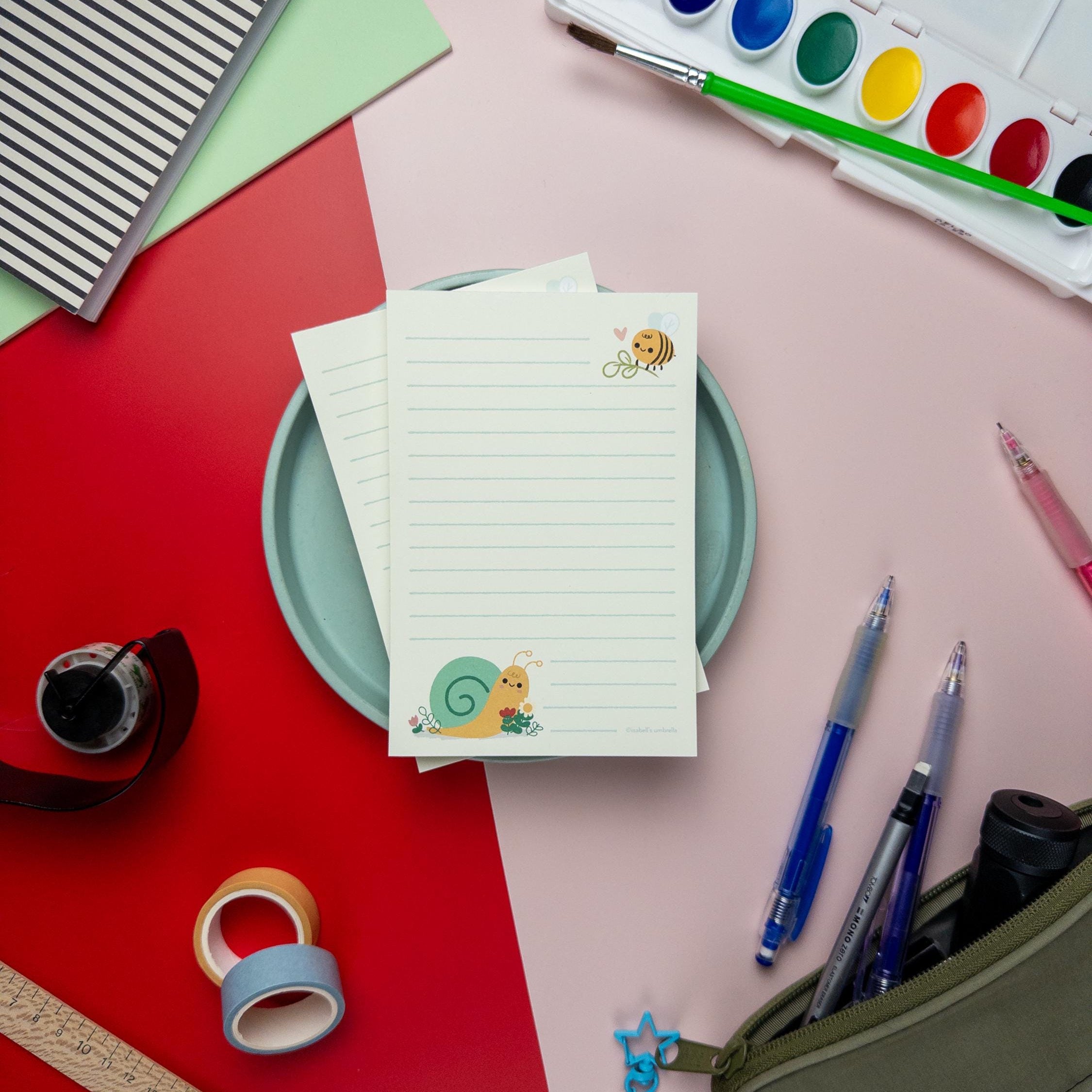 Flat lay of the snail and bee-themed notepad from Isabell’s Umbrella on a colorful desk setup. Surrounding items include watercolor paints, paintbrush, pens, washi tape, a ruler, and a pencil case, creating a cozy stationery scene.