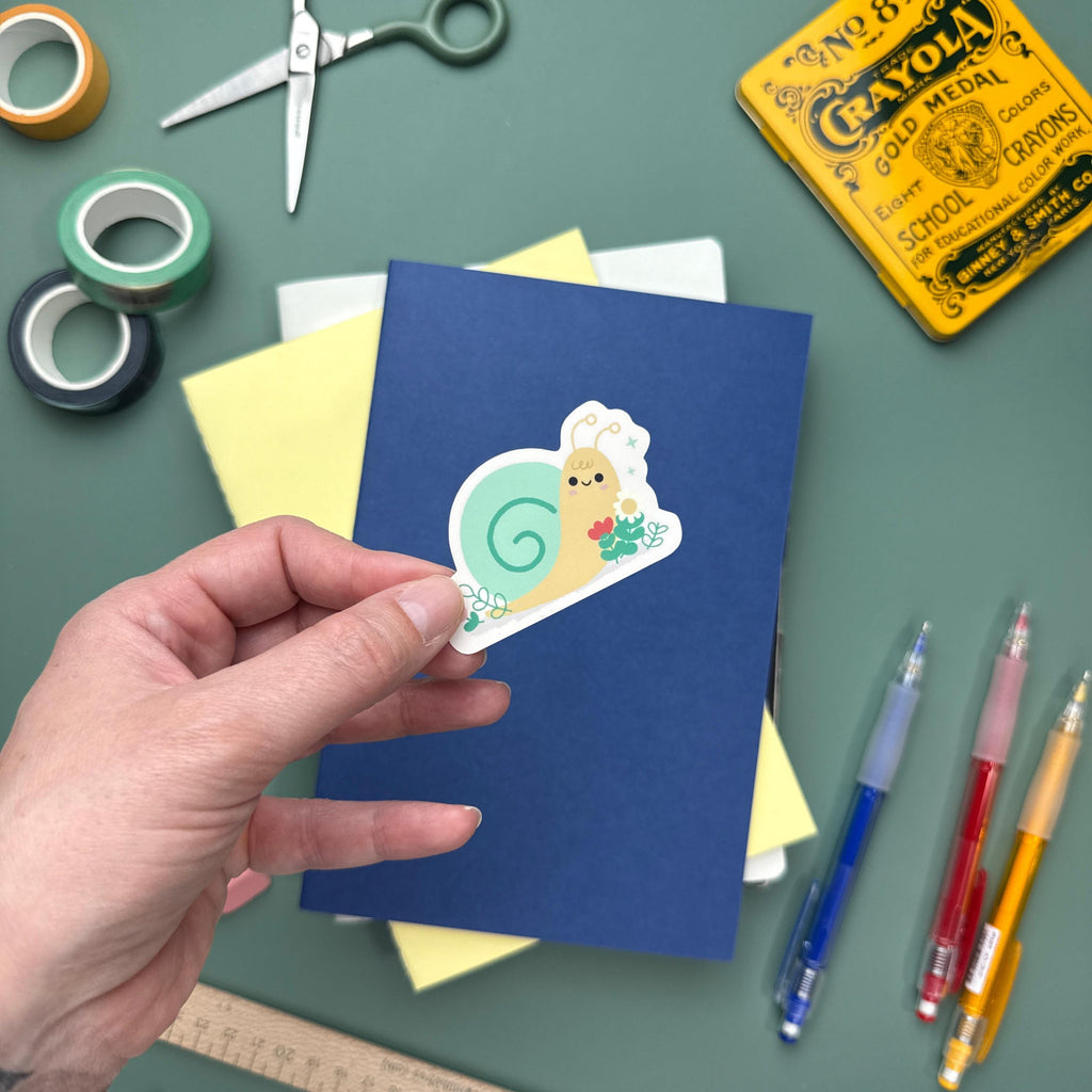 Hand holding a kawaii snail vinyl sticker featuring a sweet snail surrounded by flowers. Surrounded by colorful stationery including washi tape, scissors, and notebooks—ideal for journals, planners, scrapbooking.
