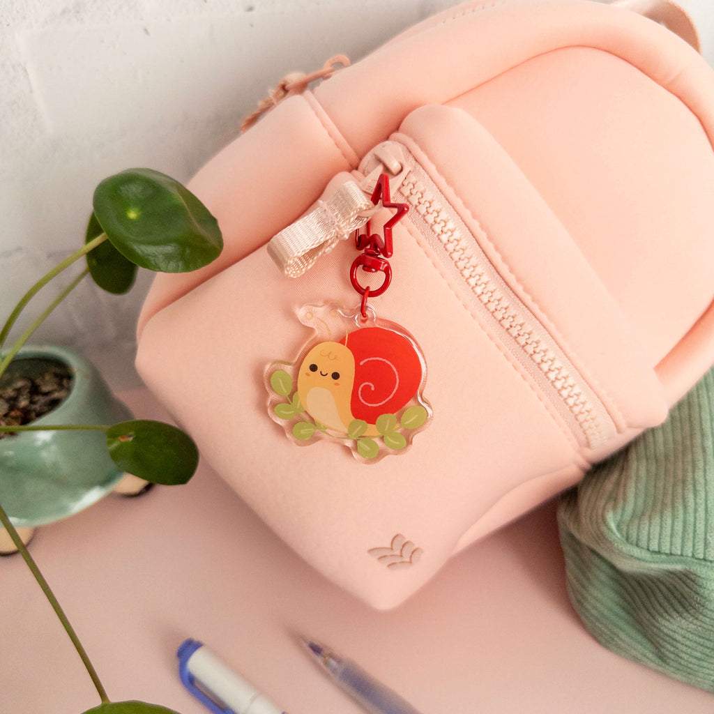 Small pink backpack  with a cute charm featuring a snail, surrounded by greenery.