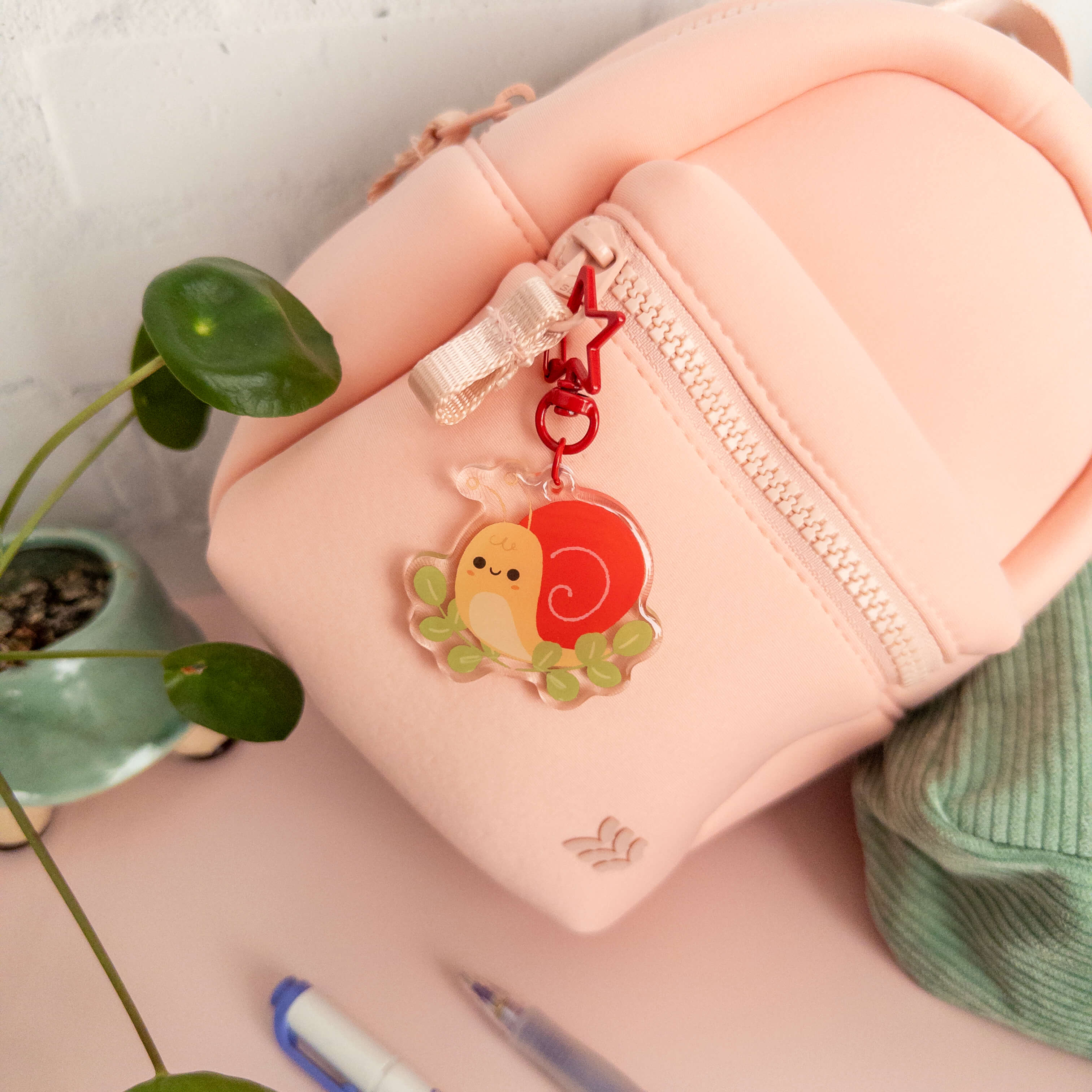 Small pink backpack  with a cute charm featuring a snail, surrounded by greenery.
