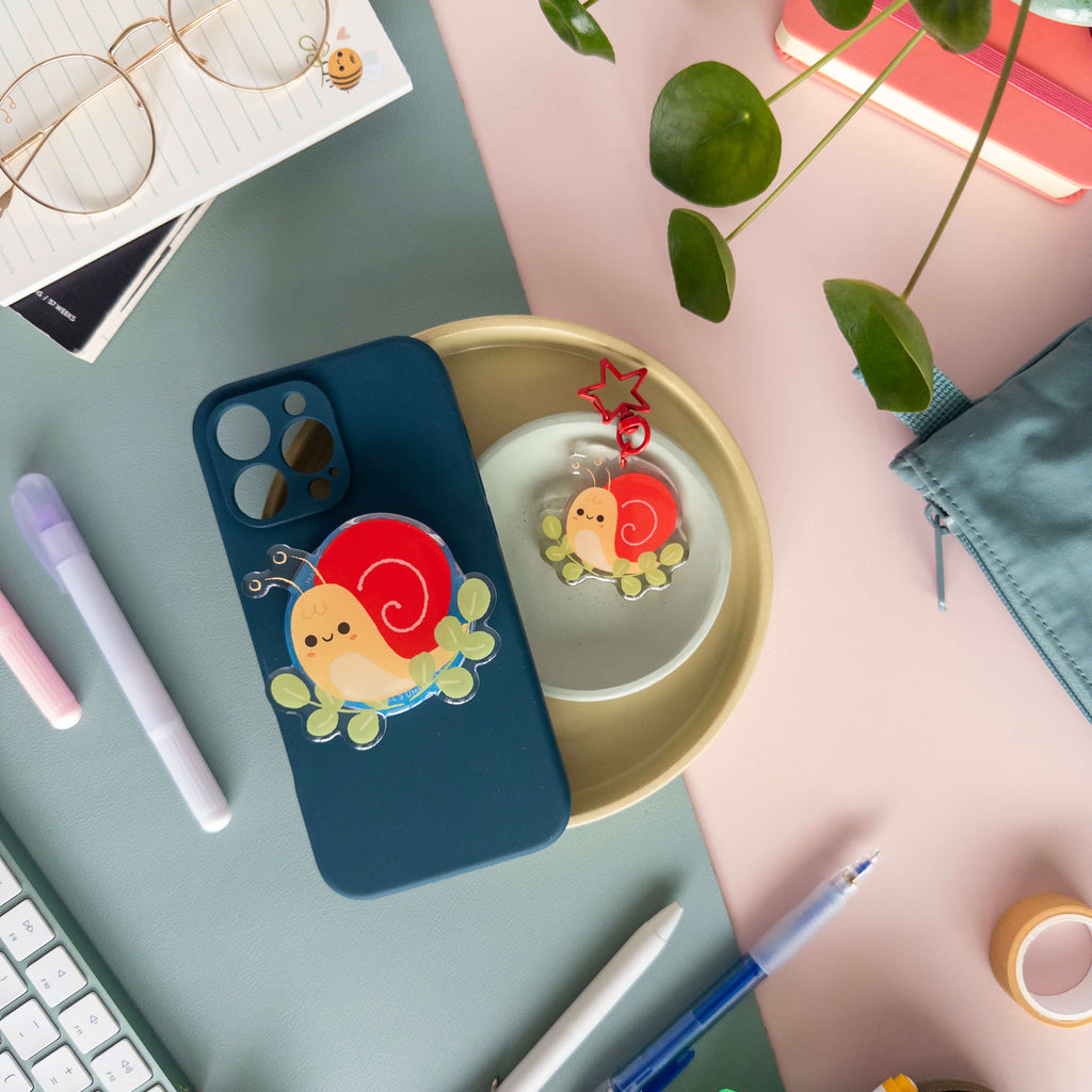 Phone case with yellow snail magnetic grip next to a snail acrylic keychain in a bowl on a desk with stationery items