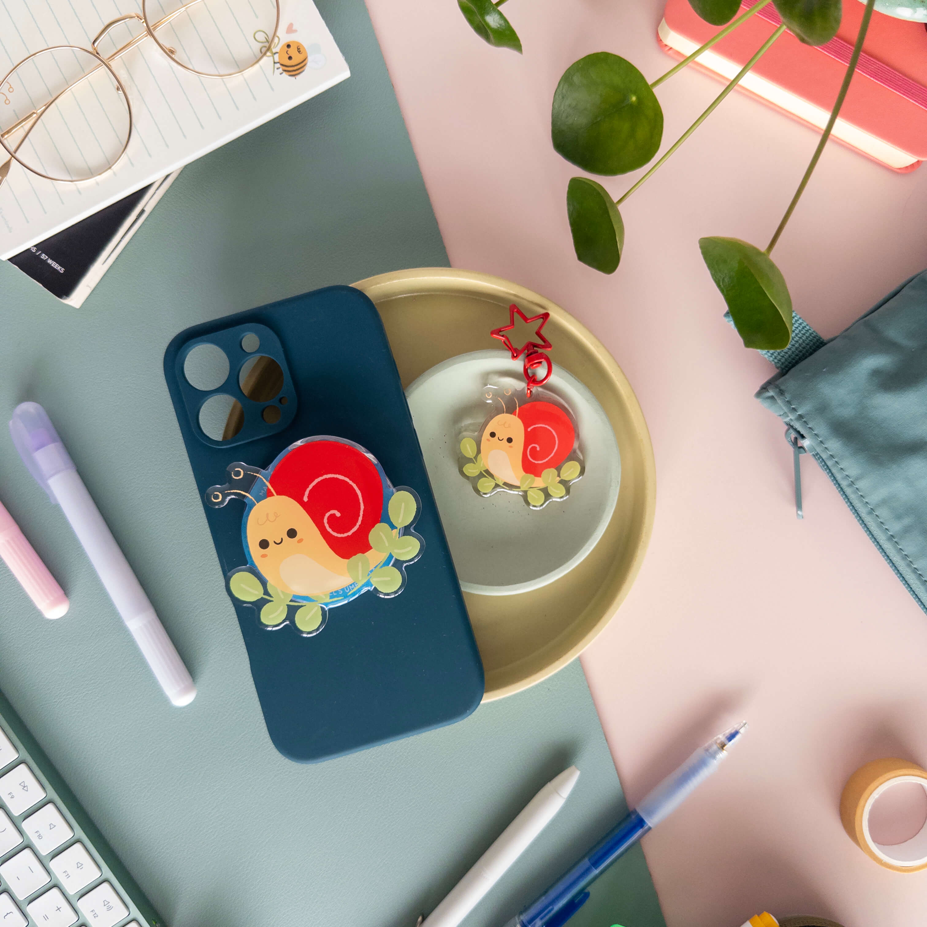 Phone case with yellow snail magnetic grip next to a snail acrylic keychain in a bowl on a desk with stationery items