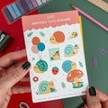 Hands holding a kawaii snail-themed vinyl sticker sheet featuring cute snails from Isabell’s Umbrella on a colorful desk setup.