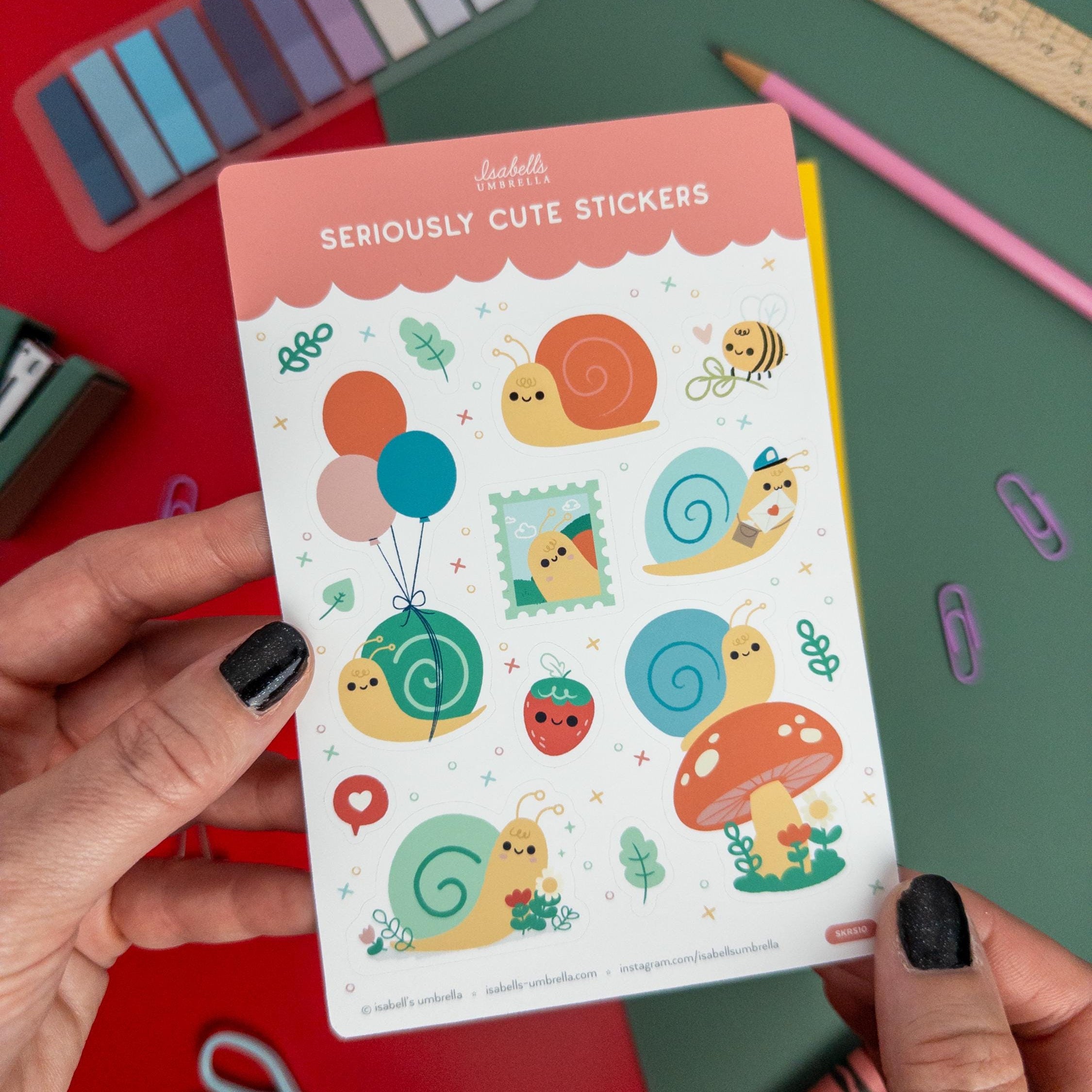 Hands holding a kawaii snail-themed vinyl sticker sheet featuring cute snails from Isabell’s Umbrella on a colorful desk setup.