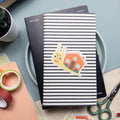 An illustrated snail with house as a shell vinyl sticker sitting atop a stack of notebooks with office supplies around.