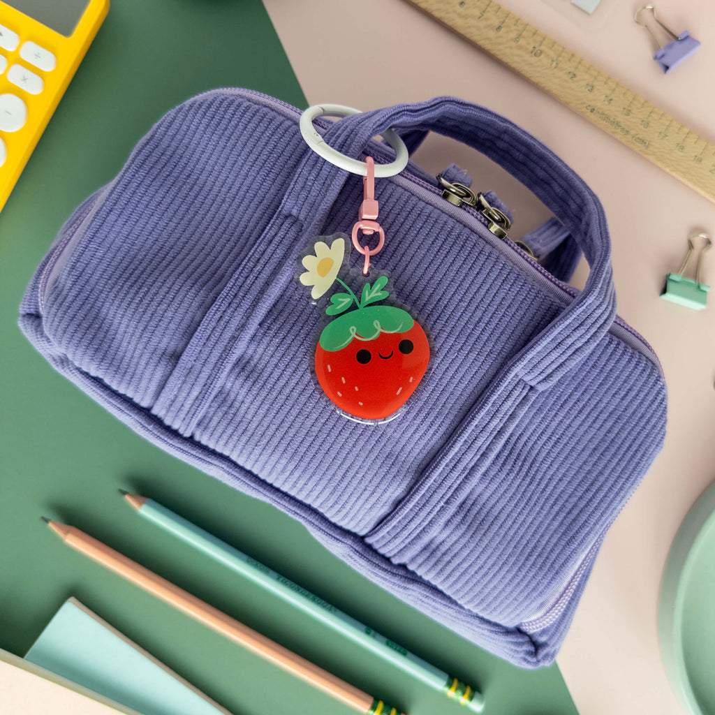 Purple corduroy pouch with a strawberry keychain on a desk with stationery items.