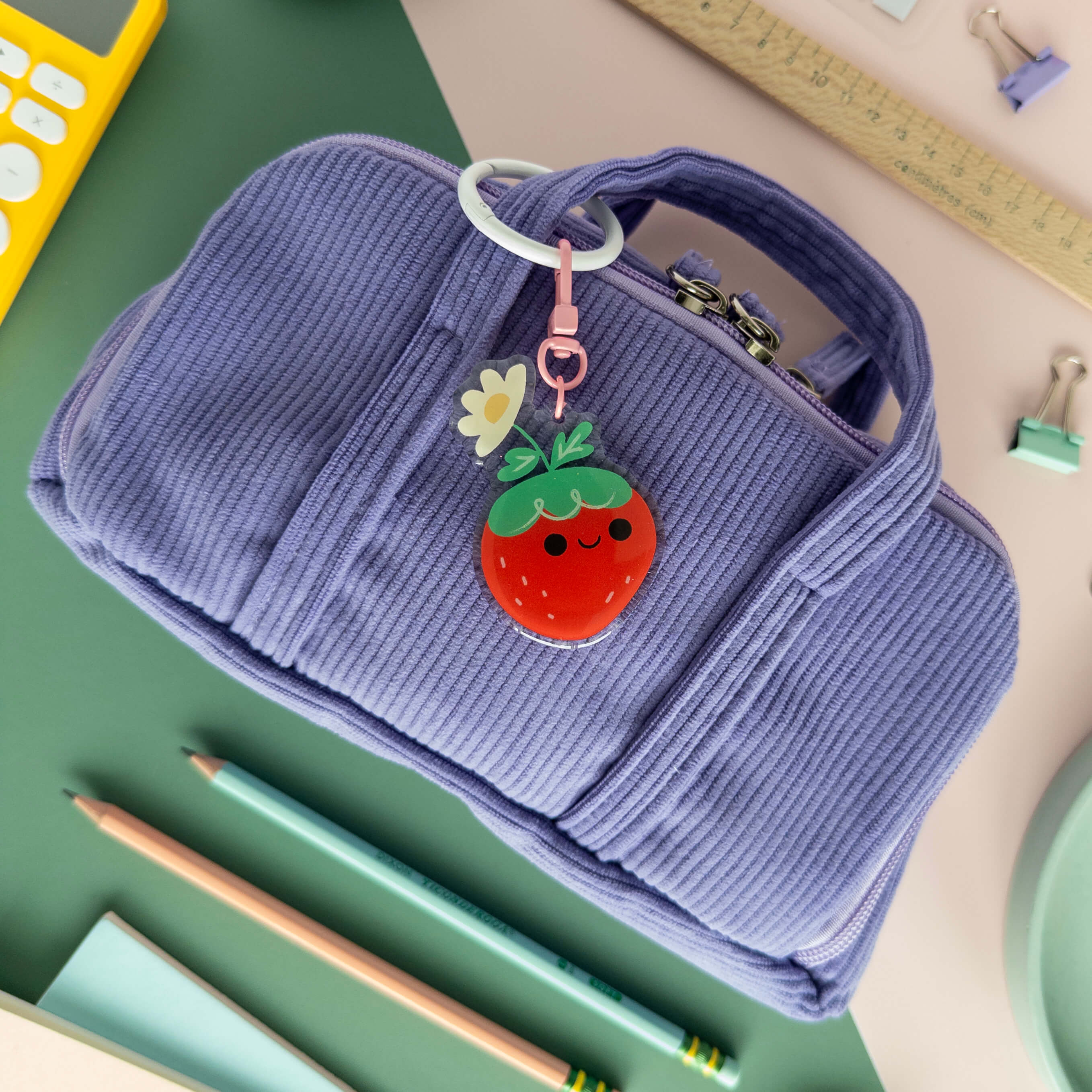 Purple corduroy pouch with a strawberry keychain on a desk with stationery items.