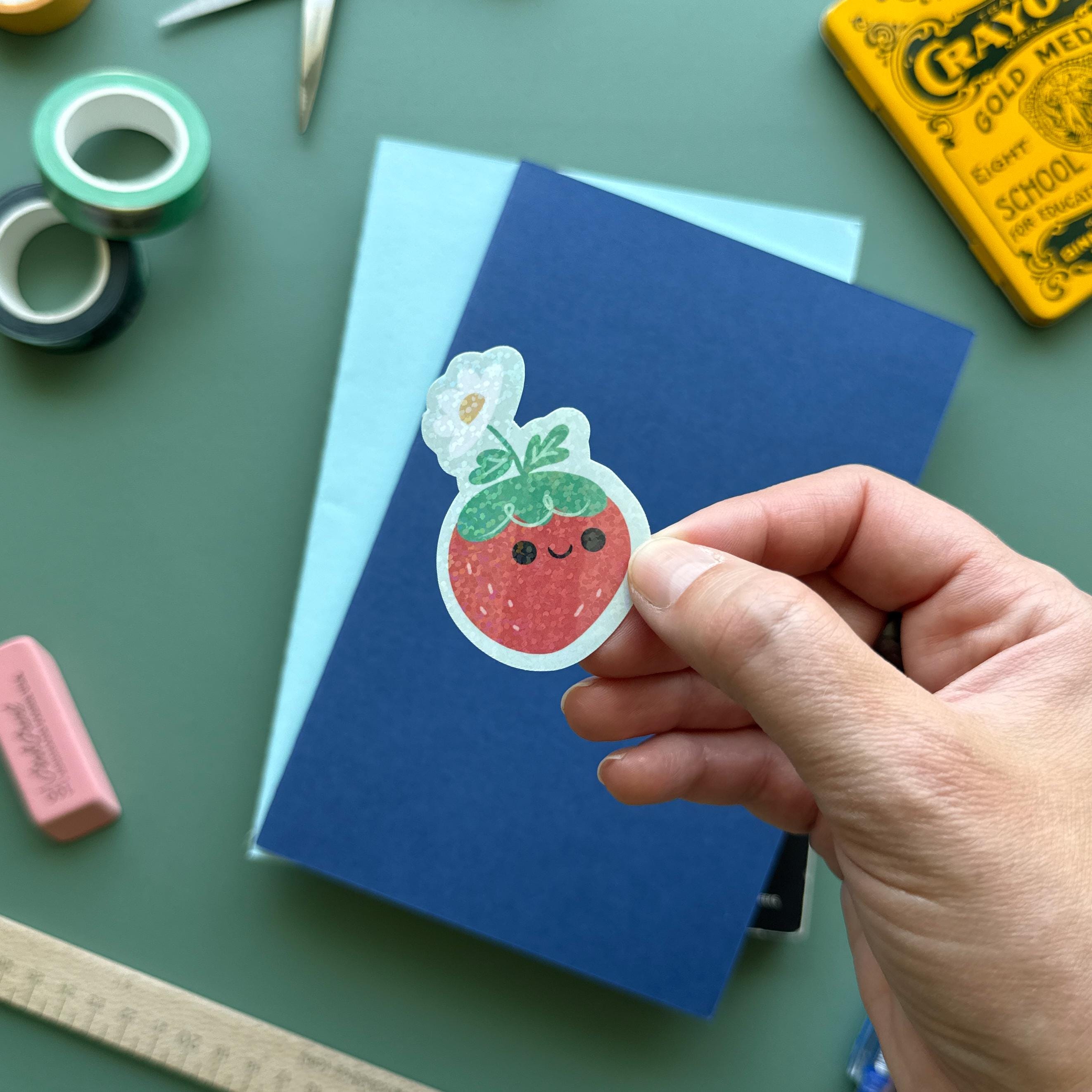 Hand holding a kawaii strawberry vinyl sticker with a flower hat. Surrounded by colorful stationery including washi tape, scissors and a notebook—ideal for journals, planners, and scrapbooking.