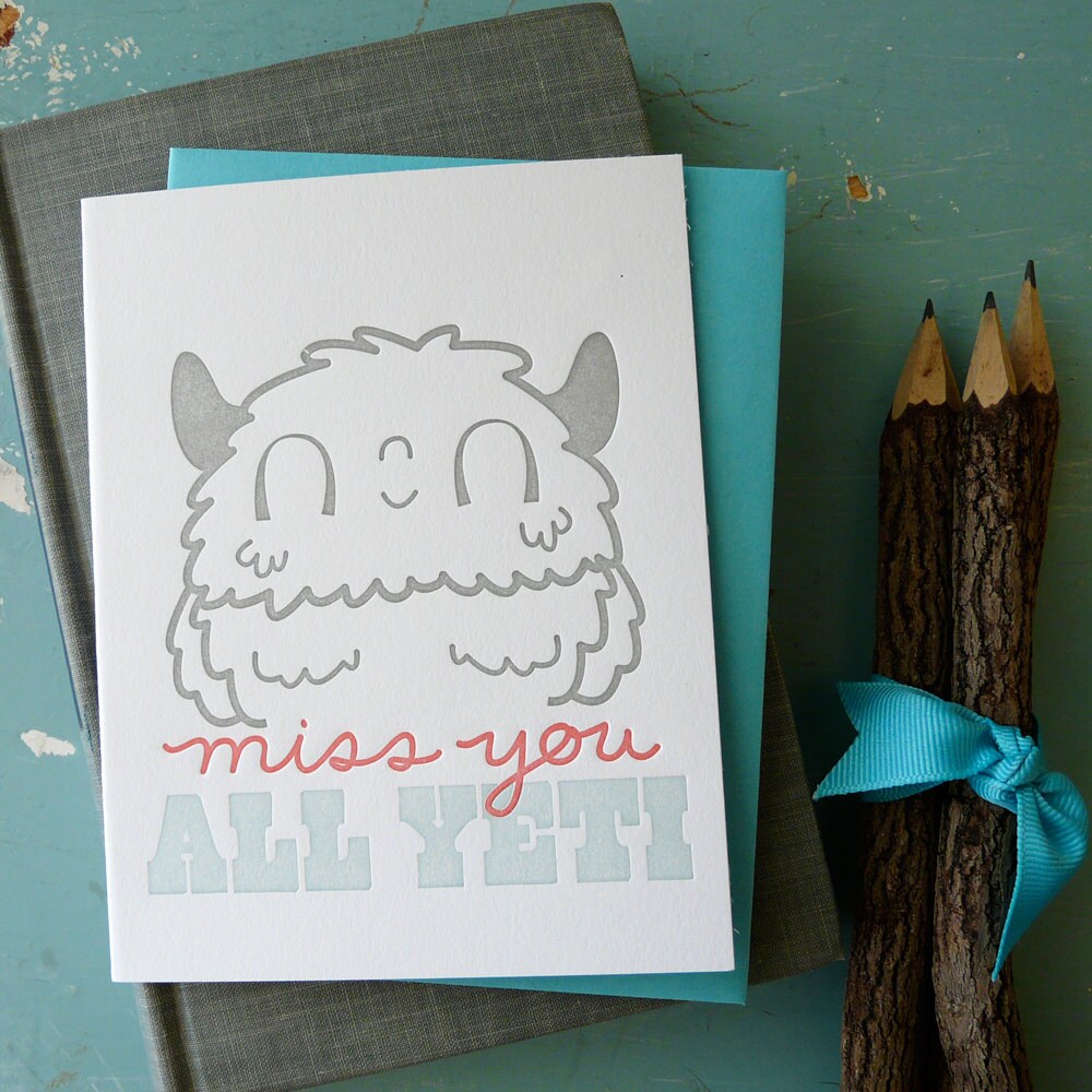 A cute illustrated letterpress card featuring a friendly yeti with “Miss You All” message sitting on top of a book with a set of pencils beside it.