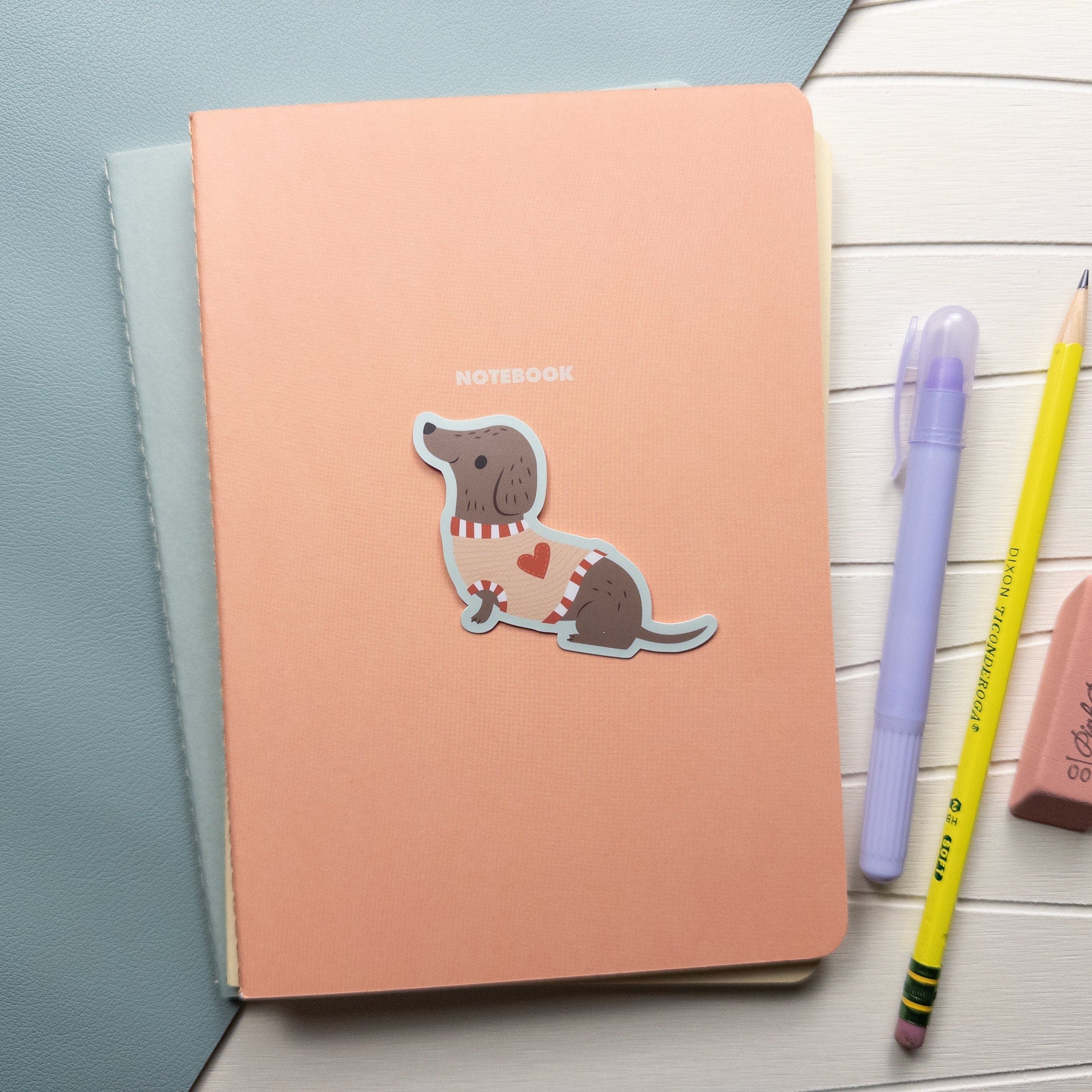 An illustrated vinyl sticker of a dachshund wearing a sweater sitting on top of a notebook with a marker, pencil, and erasure off to the right.