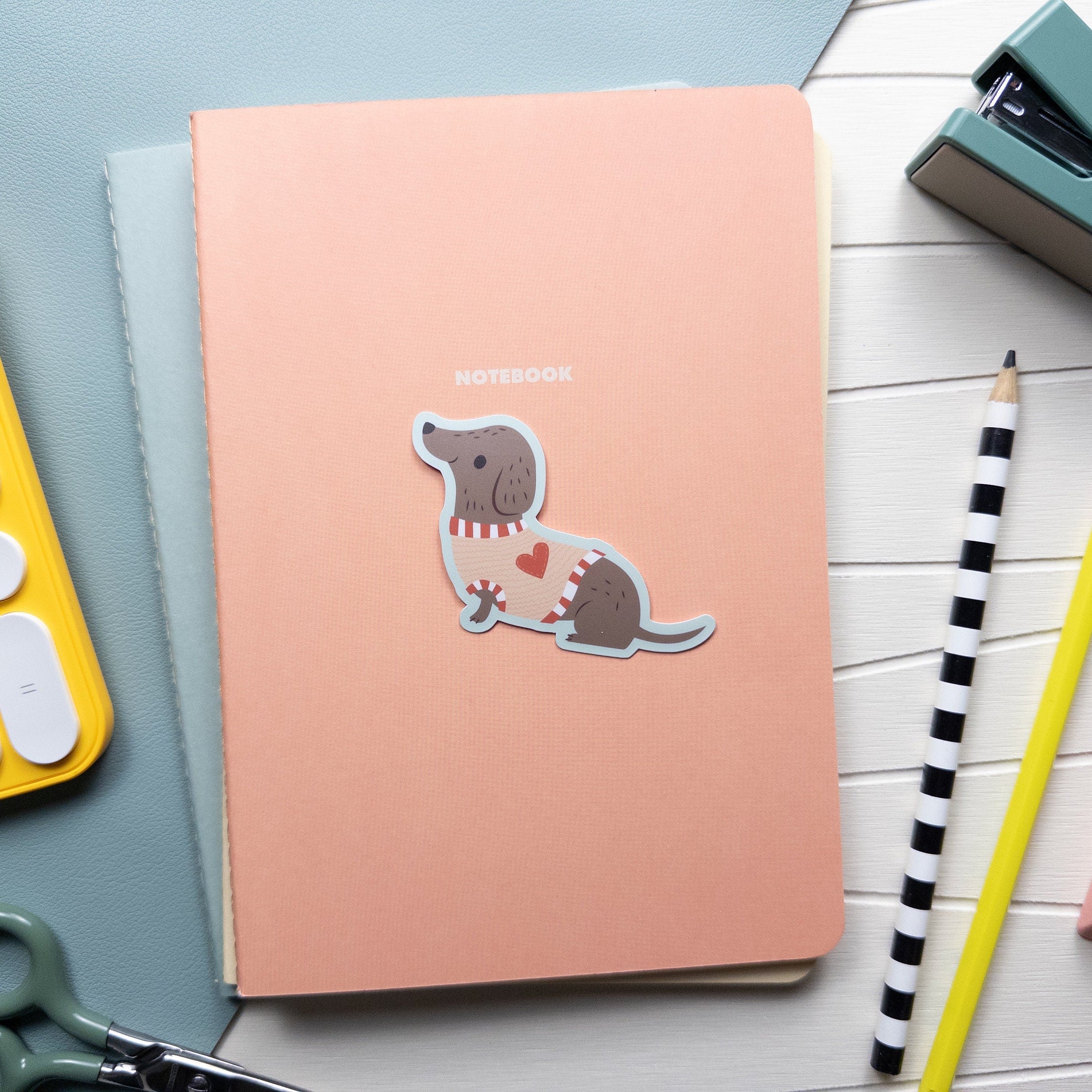 An illustrated waterproof vinyl sticker of a dachshund wearing a heart sweater sitting atop a pile of notebooks surrounded by office supplies.