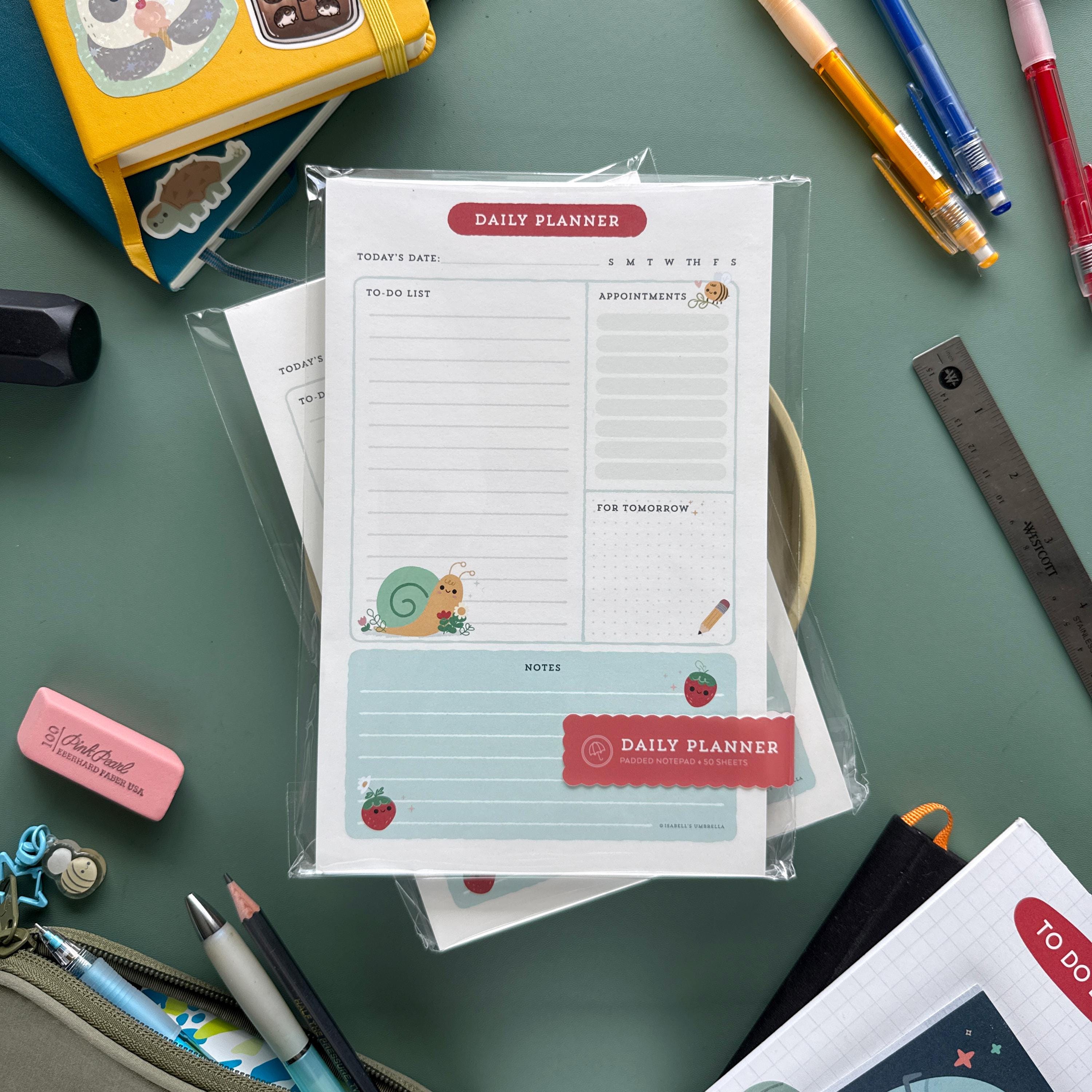 Packaged daily planner notepads with colorful insect illustrations lie on a green surface, ready for sale. Nearby are notebooks with stickers, pens, a ruler, an eraser, and other office supplies, creating a creative and organized workspace vibe.