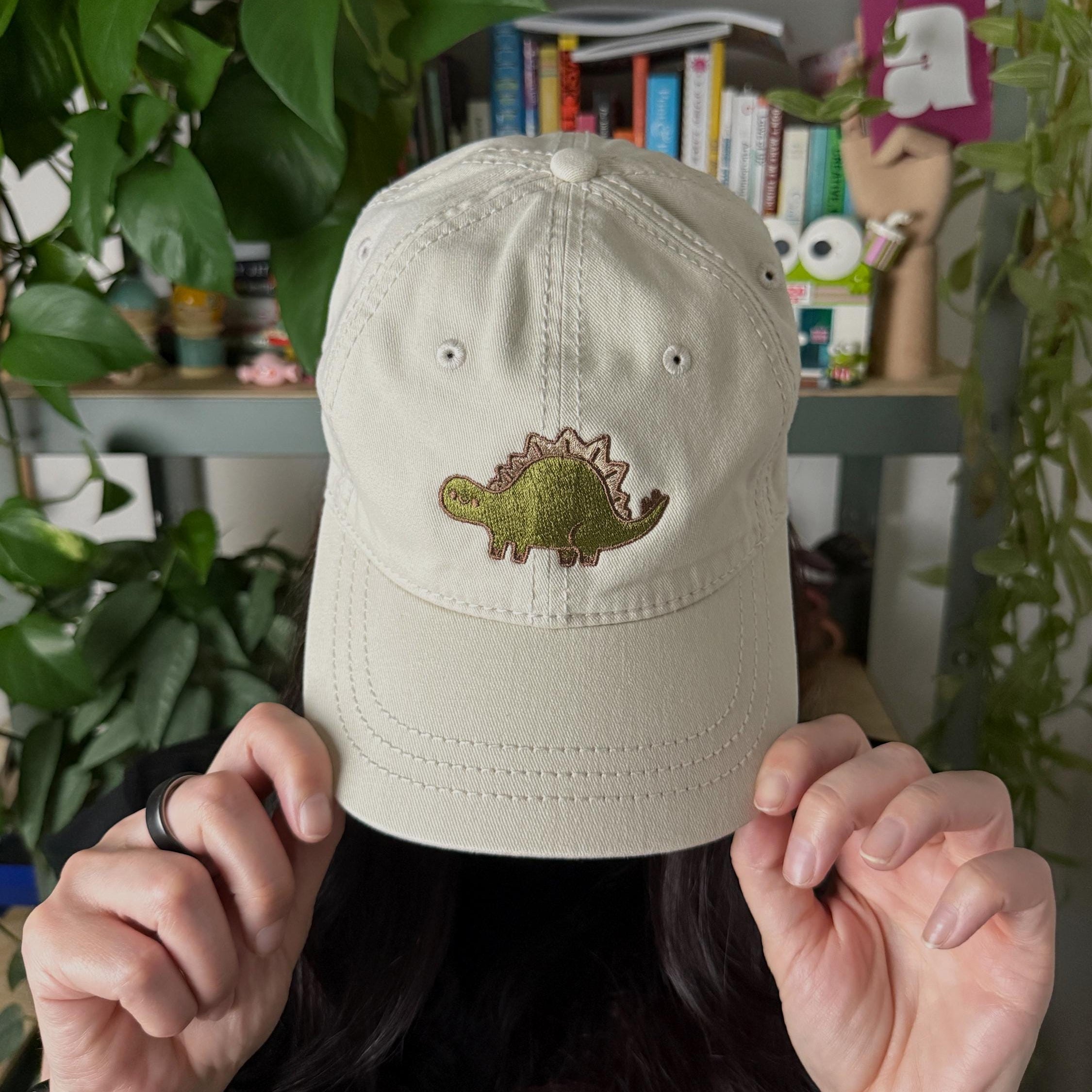 Woman wearing a cream cotton twill dad hat with embroidered green stegosaurus design pulled down low on her face.