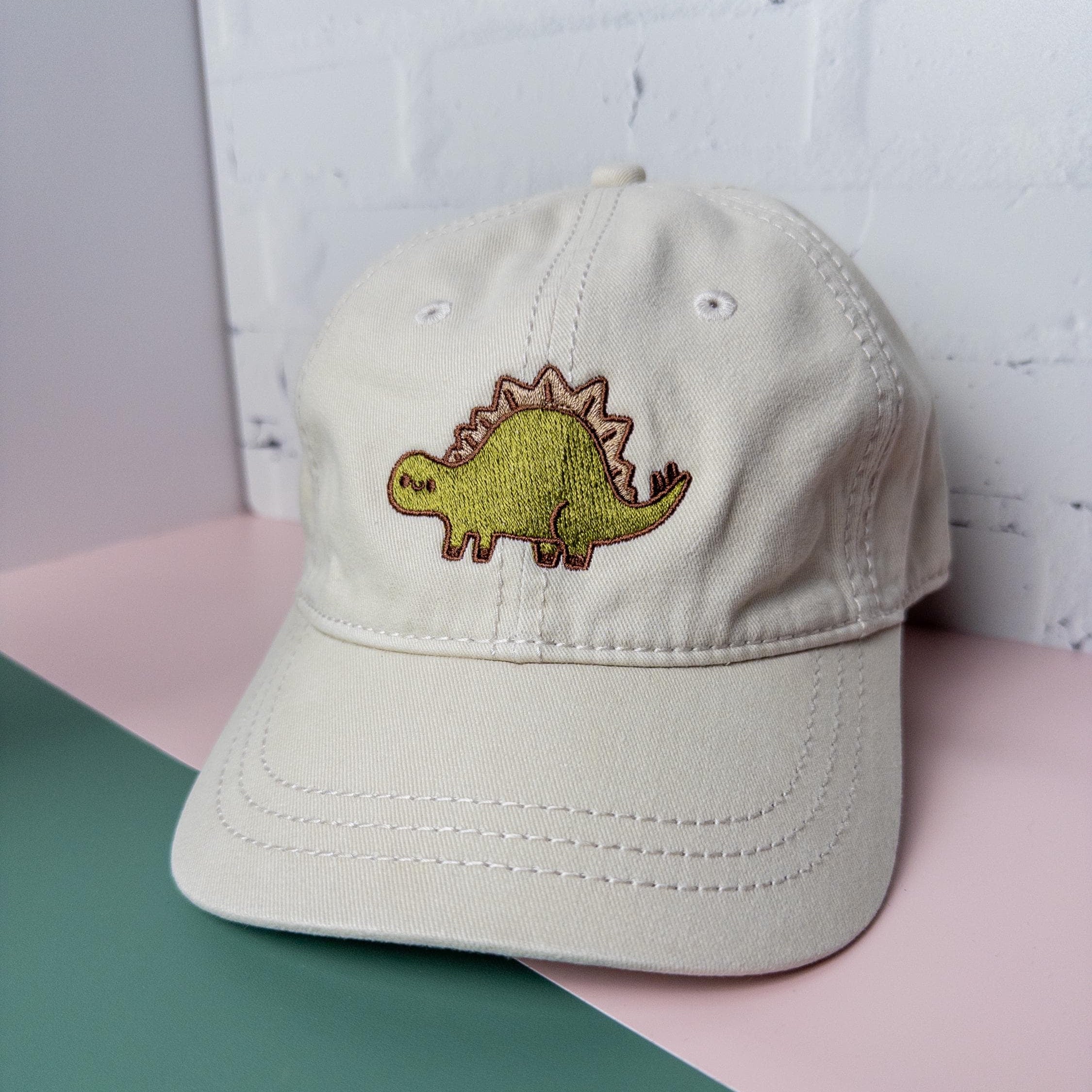 Cream cotton twill dad hat with embroidered green stegosaurus design pictured on top of a pink and green table top.