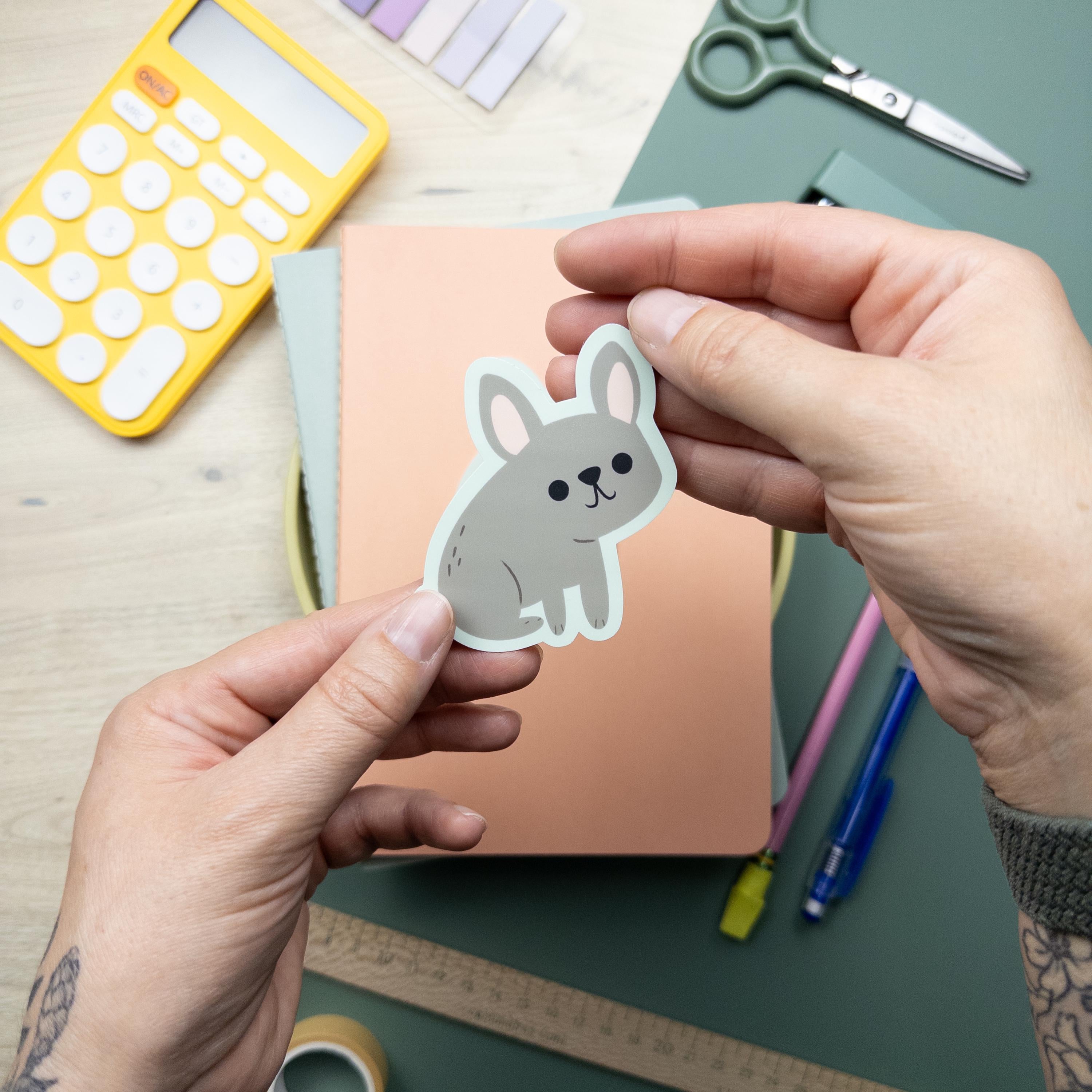 Hand holding a cute illustrated French Bulldog vinyl sticker, waterproof Frenchie dog decal for laptops, water bottles, and journals, durable matte vinyl design.