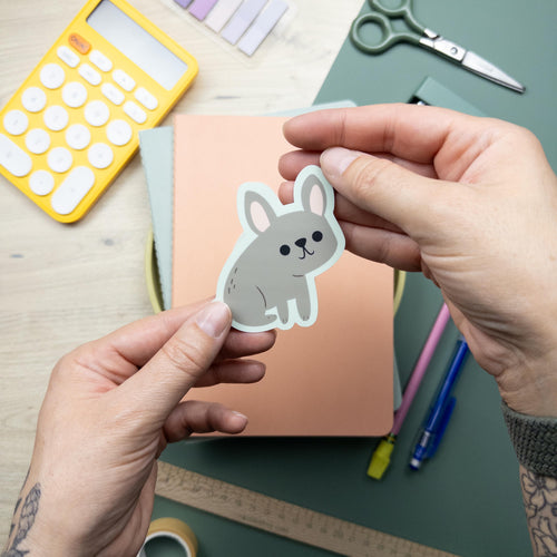Hand holding a cute illustrated French Bulldog vinyl sticker, waterproof Frenchie dog decal for laptops, water bottles, and journals, durable matte vinyl design.