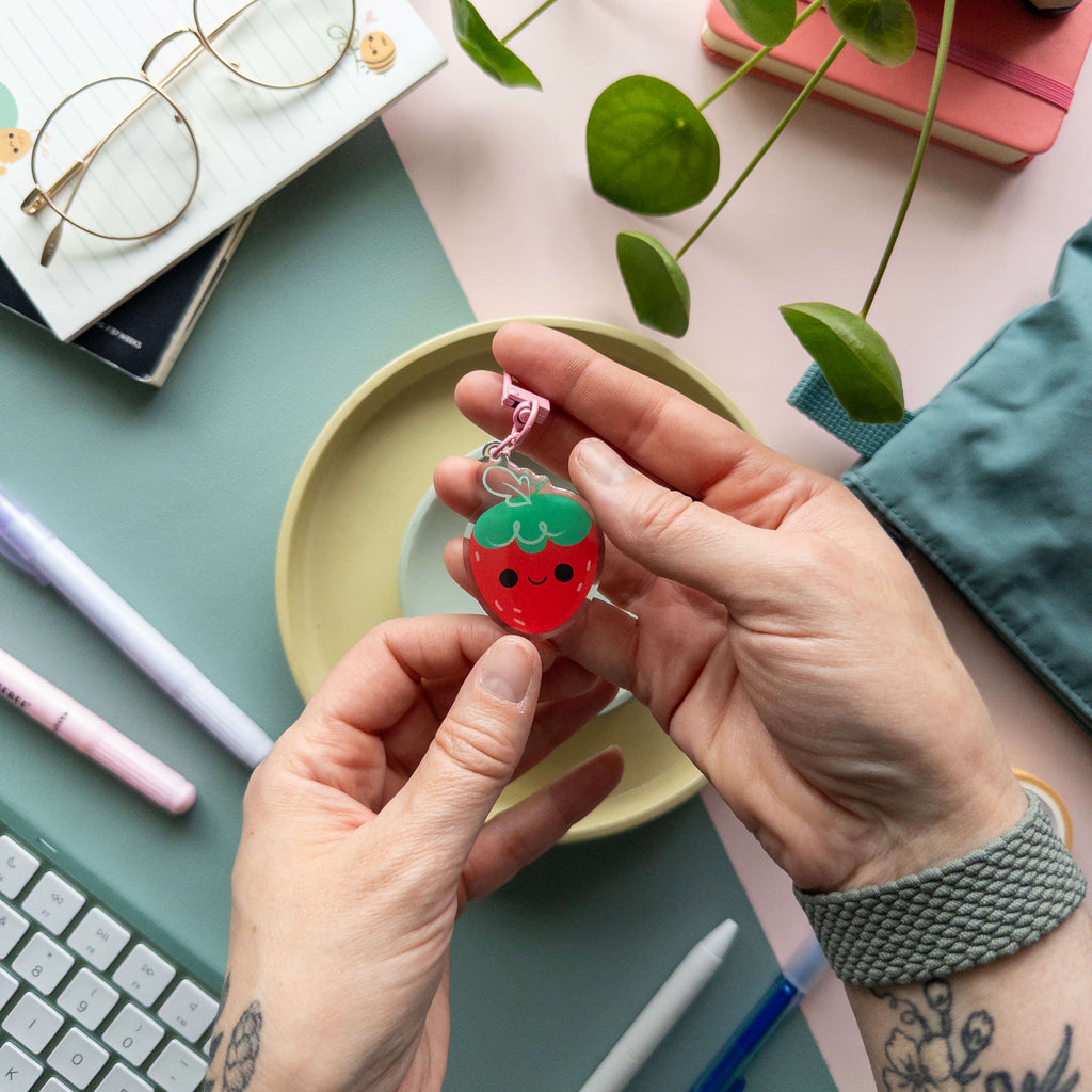 Person holding a strawberry-shaped keychain on a desk with stationery items and a plant.