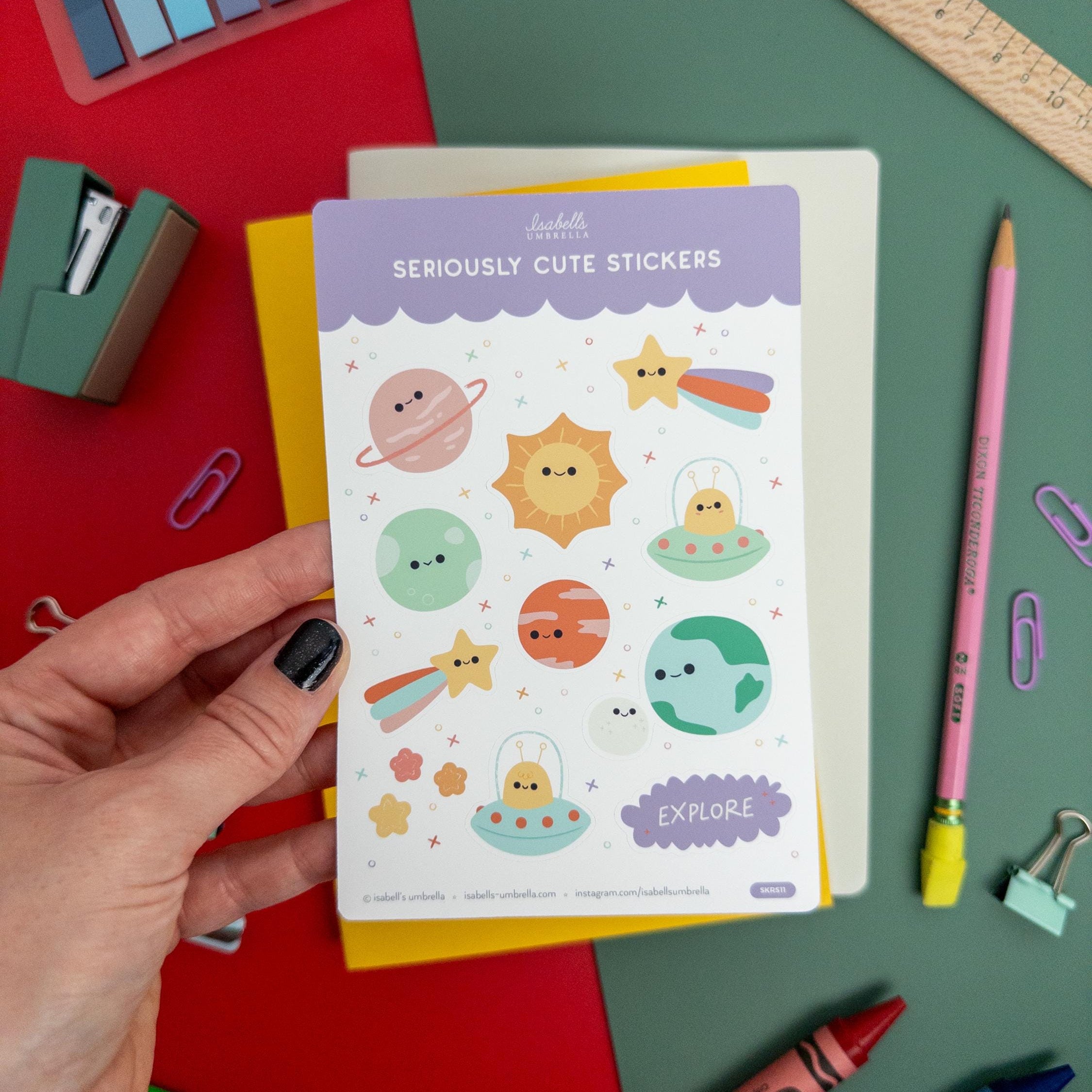 Hand holding a kawaii planet-themed vinyl sticker sheet featuring cute suns, planets, and stars from Isabell’s Umbrella on a colorful desk setup.