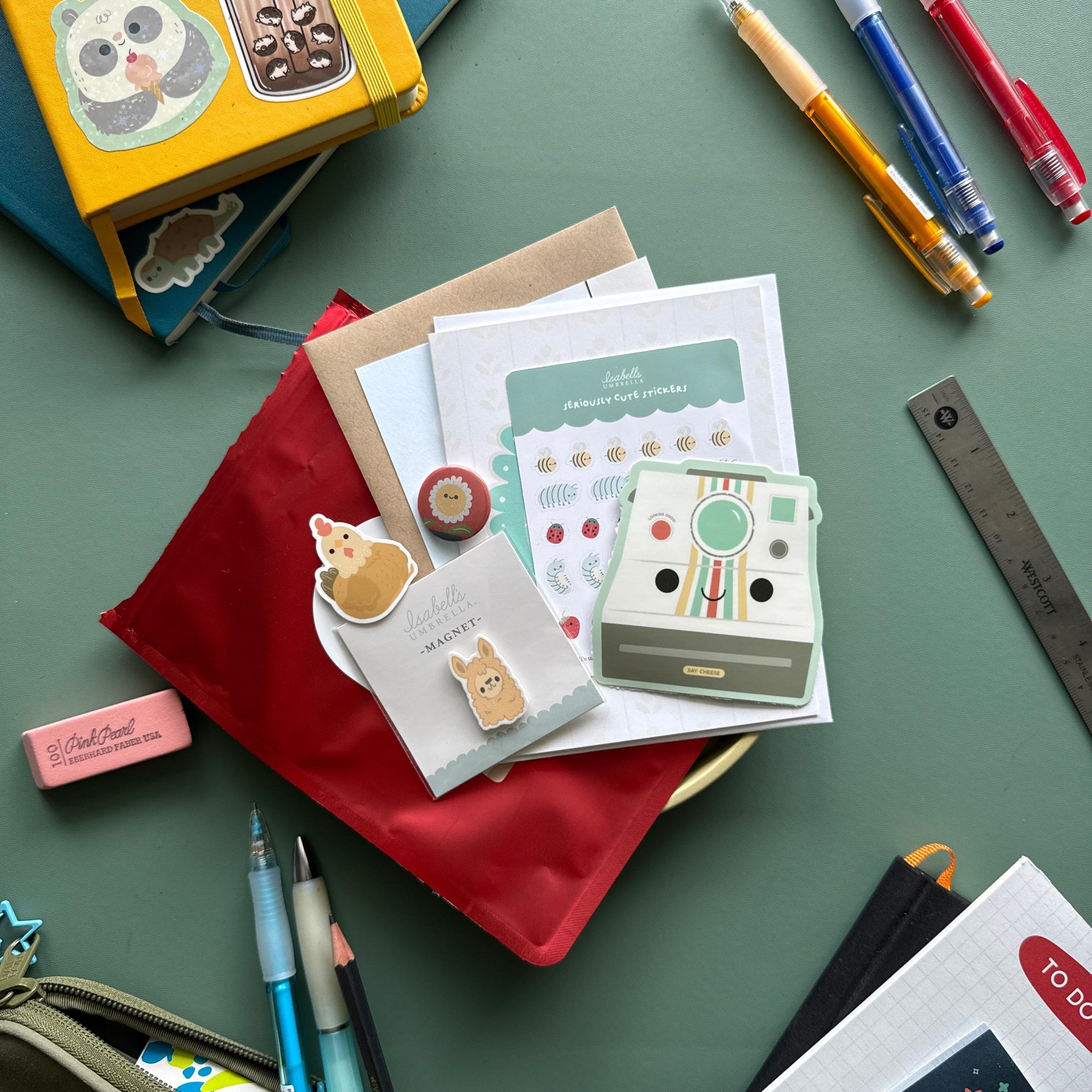 collection of products including a magnet, button, stickers, and note cards pictured on top of a red bag surrounded by a variety of office and art supplies.
