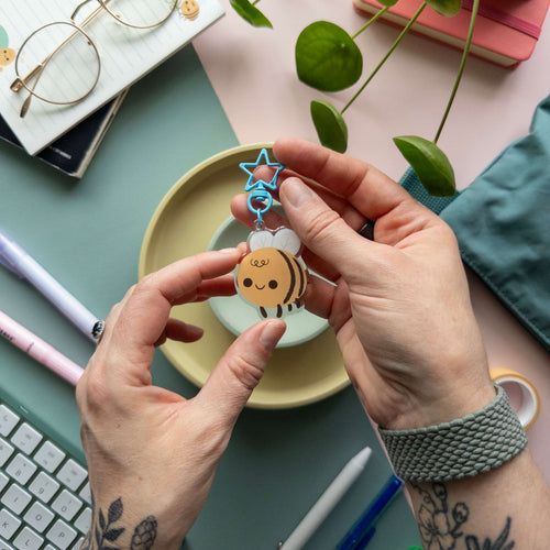 Person holding a small decorative keychain featuring a bee with blue star clasp on a desk with stationery items.